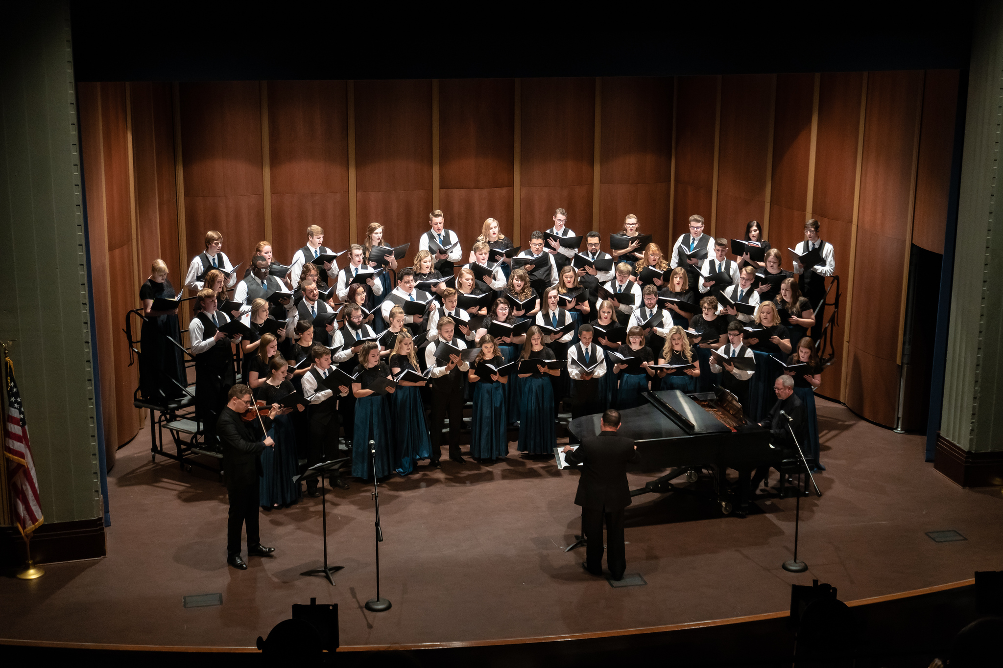 Large mixed choir performing on stage with a conductor, a violinist, and a pianist, all dressed formally, with wooden paneling backdrop.