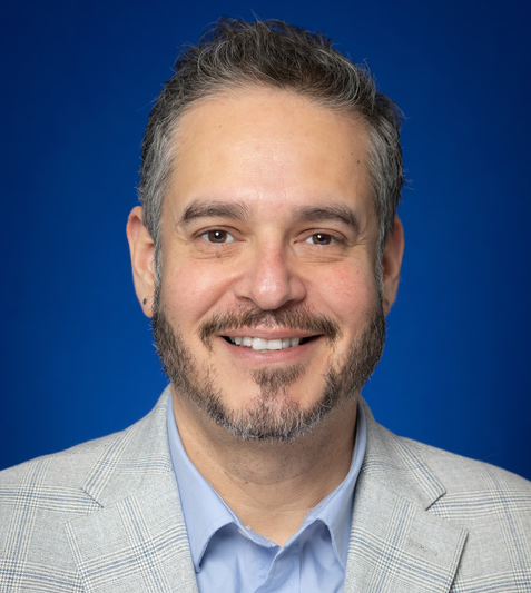 Man with short dark hair and beard wearing a light gray plaid blazer and light blue shirt against a blue background smiling.
