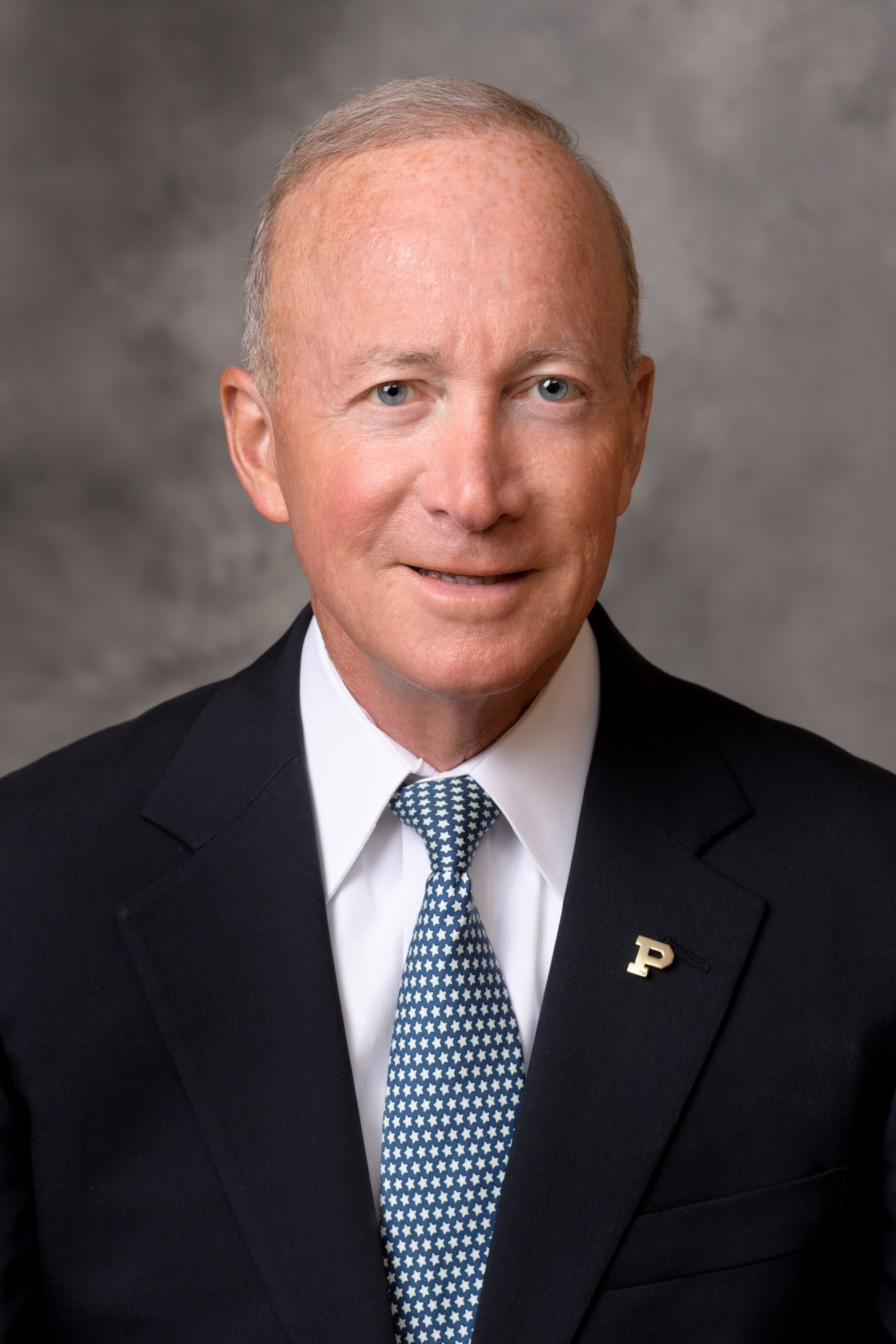 Older man with short light hair wearing a dark suit, white shirt, and blue tie with white stars, against a gray mottled background