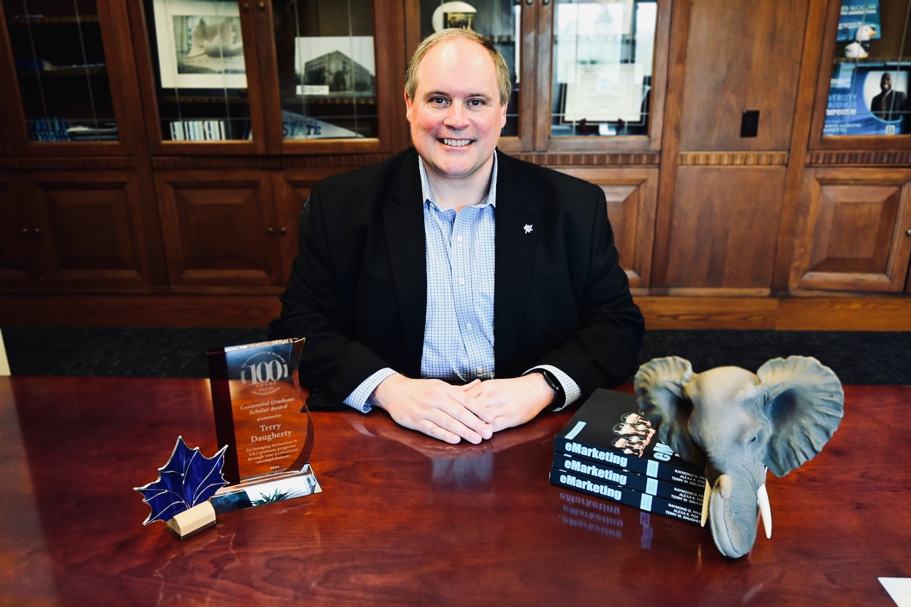 Man in black blazer and blue checkered shirt sitting at a wooden table with an award, three marketing books, and an elephant head sculpture.