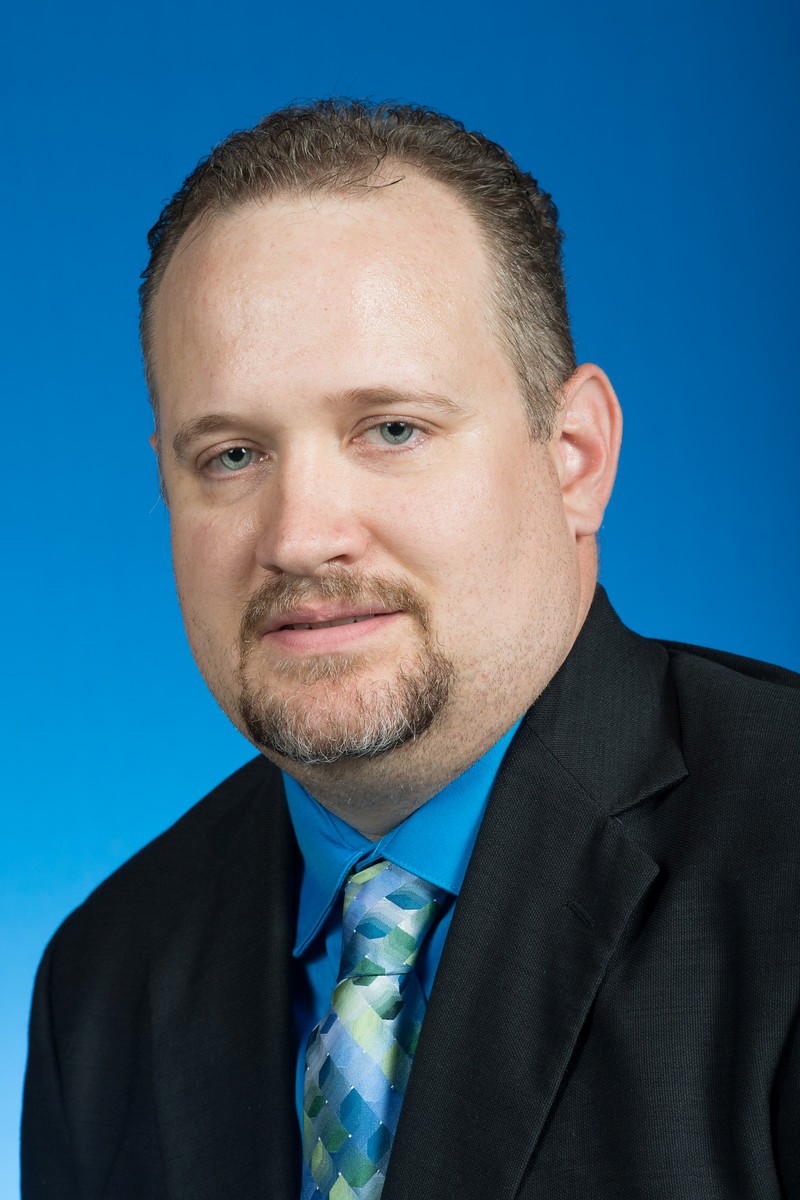 Man with short curly hair and trimmed beard wearing a black suit, blue shirt, and patterned tie, against a solid blue background.