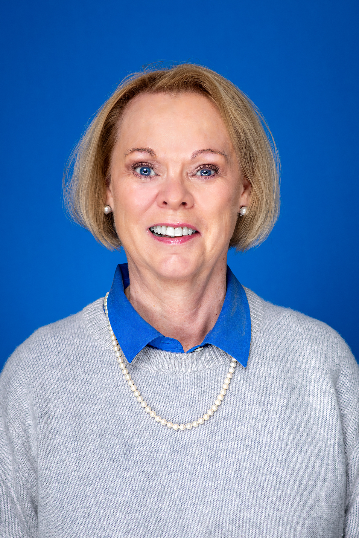Woman with short blonde hair wearing pearl earrings and necklace, a blue collared shirt under a gray sweater, smiling against a blue background.