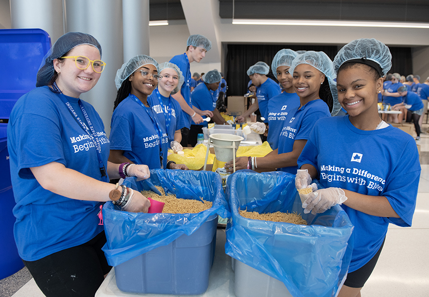 Group of young volunteers in blue shirts and hairnets packing food into large blue bins at a community service event indoors.