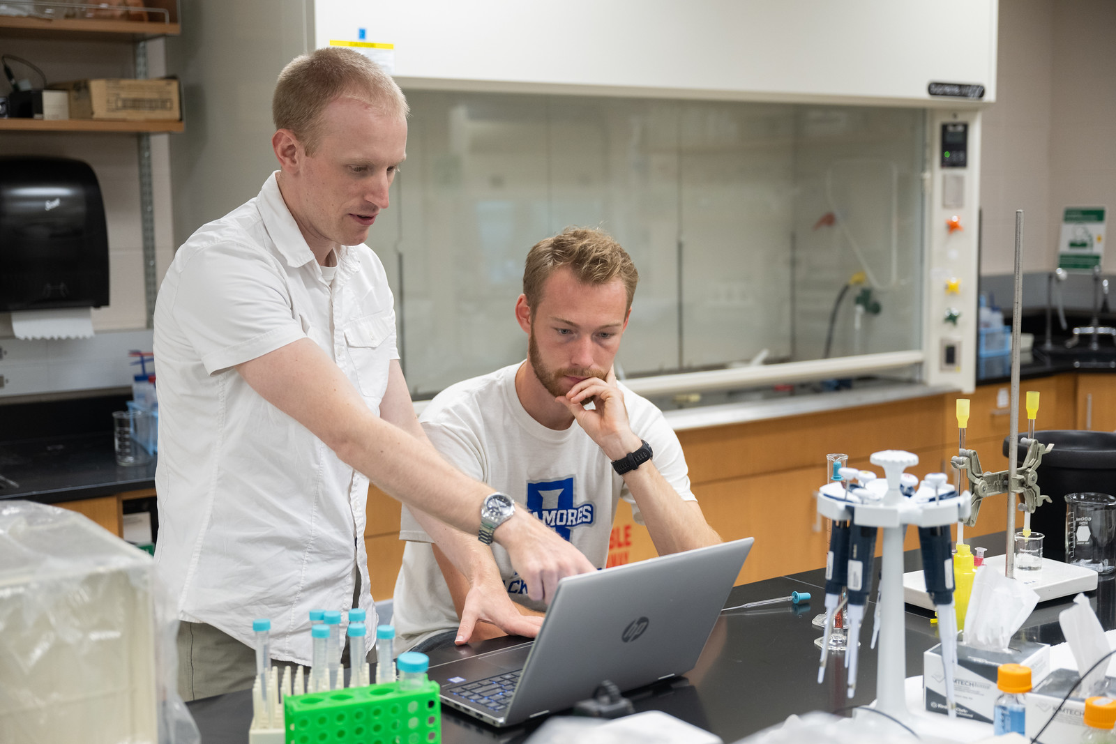 Two men in a lab, one pointing at a laptop screen while the other watches thoughtfully, surrounded by lab equipment and test tubes.