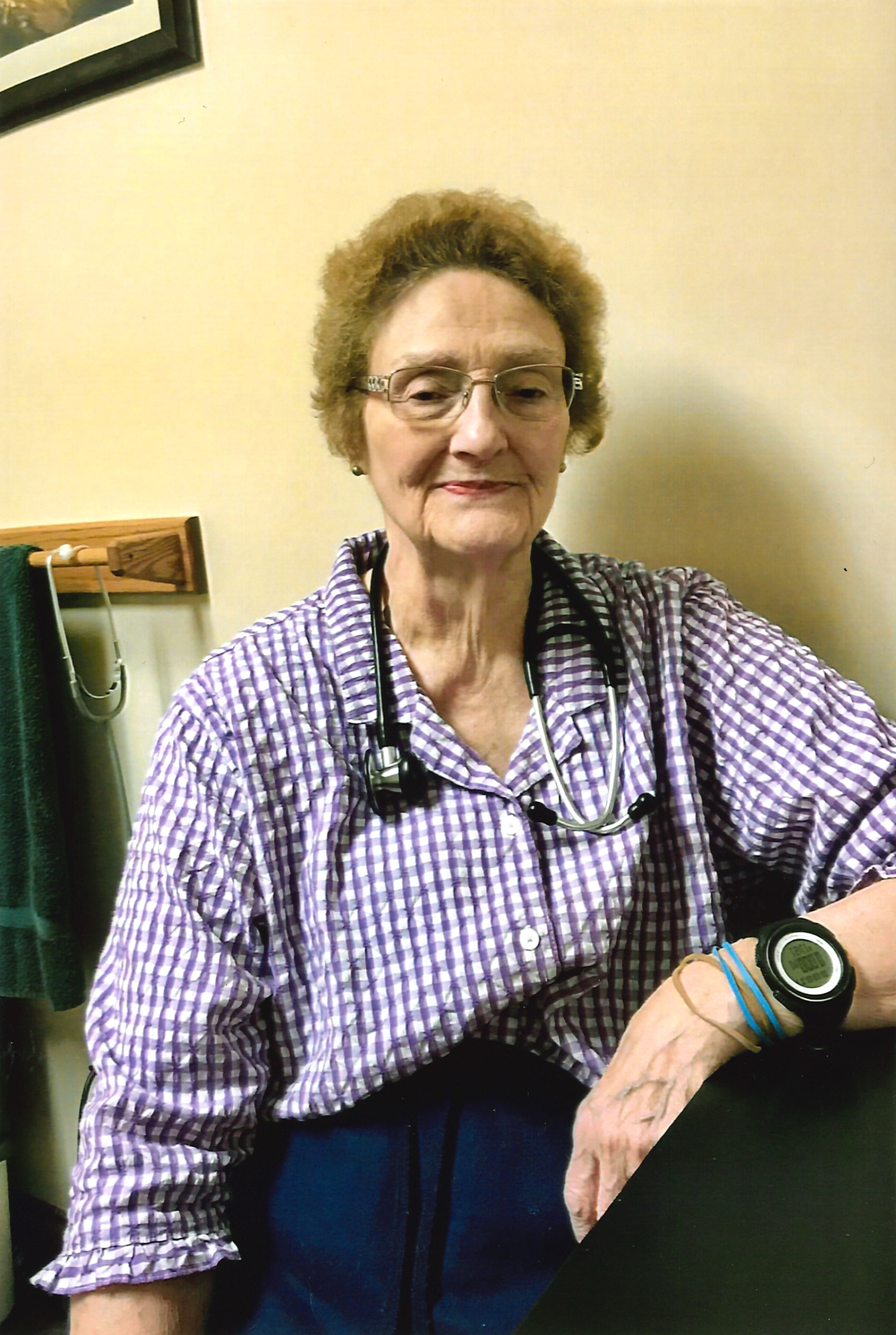 Elderly woman wearing glasses, a purple checkered shirt, and a stethoscope around her neck, seated near a towel rack with a dark green towel.