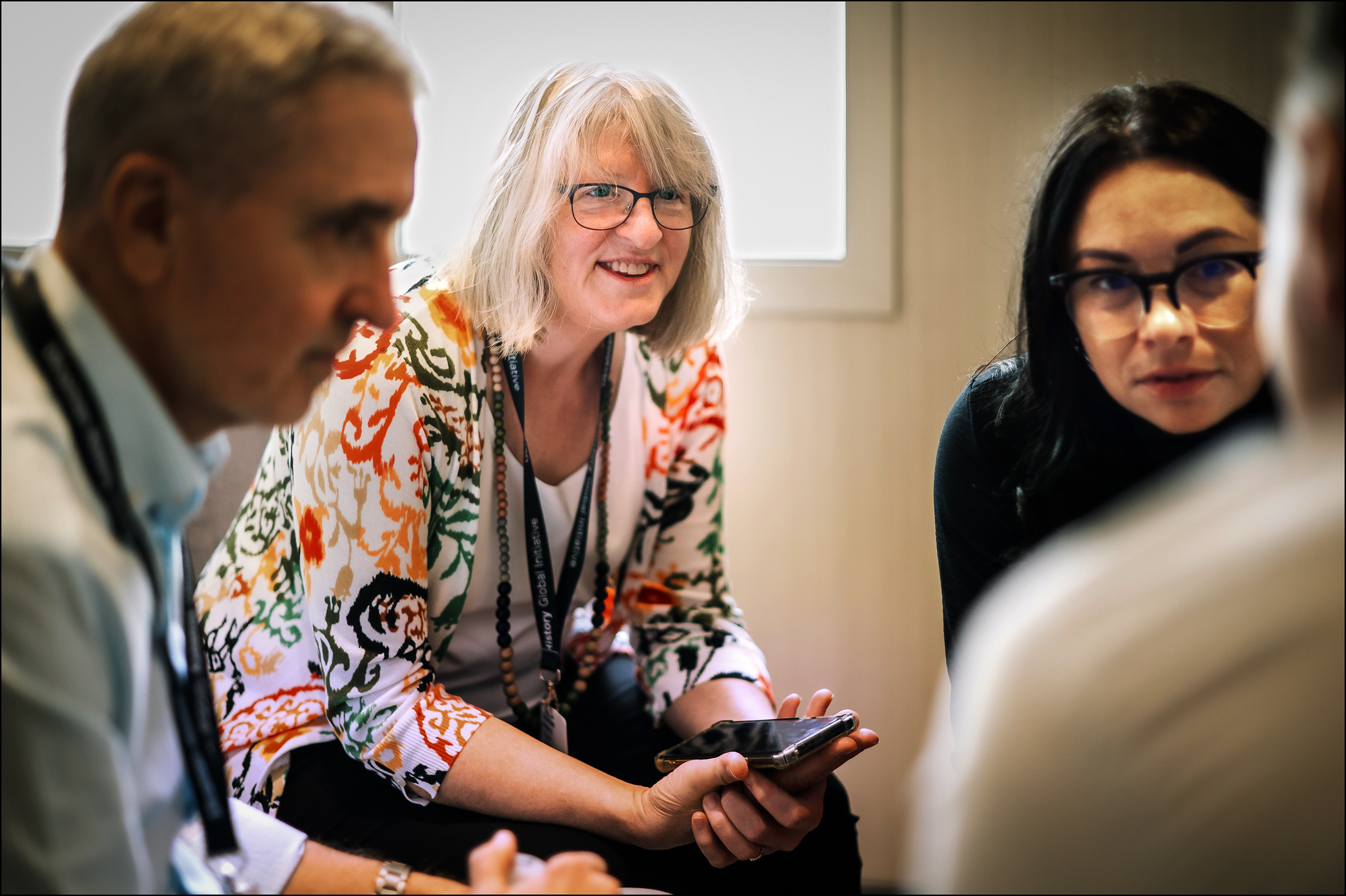 Four people engaged in discussion, one woman with glasses holding a phone and wearing a colorful patterned shirt smiling, others listening attentively.