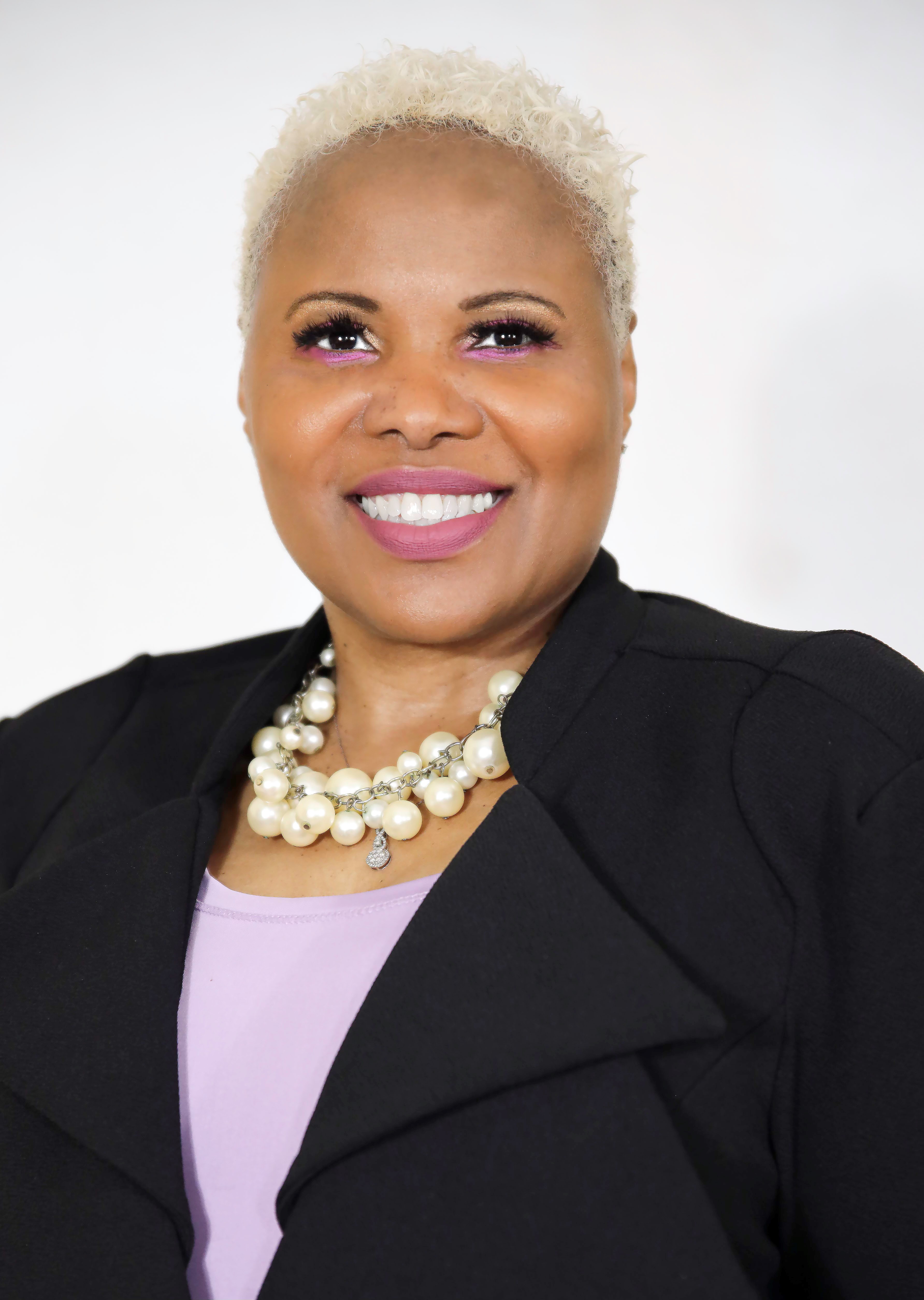 Smiling woman with short blonde hair wearing a black blazer, lavender top, and a large pearl necklace against a plain white background.