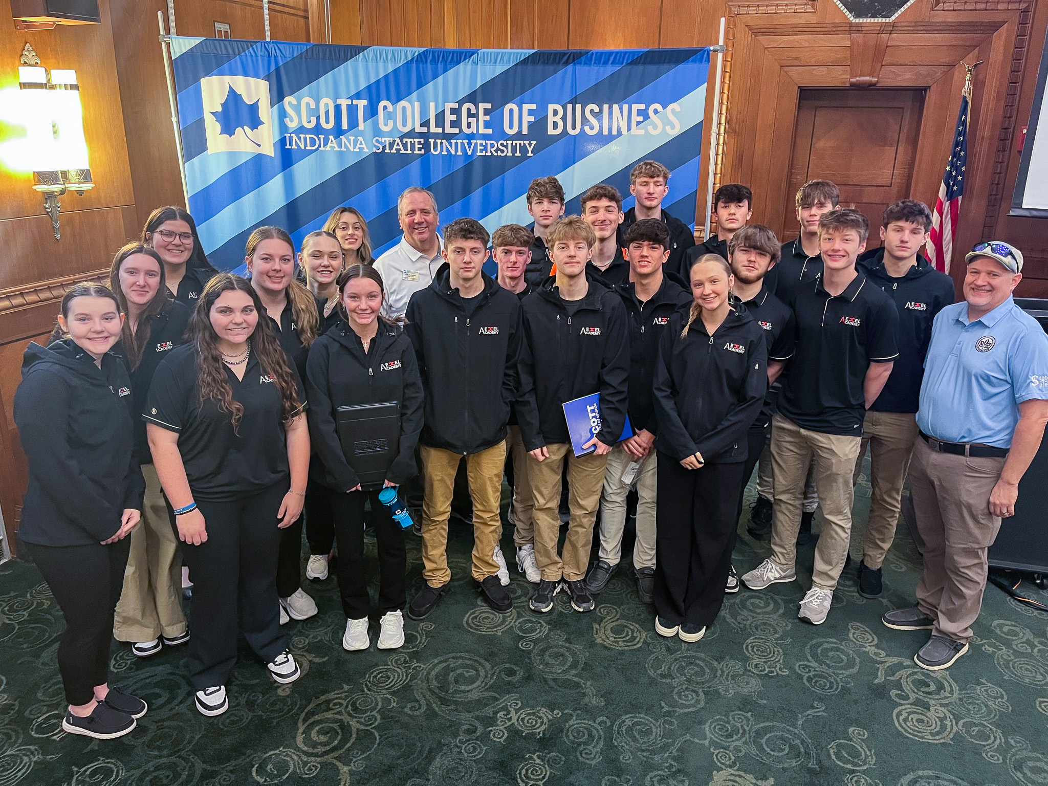 Group of students and two adults wearing matching black and blue outfits posing indoors in front of a Scott College of Business banner at Indiana State Universi