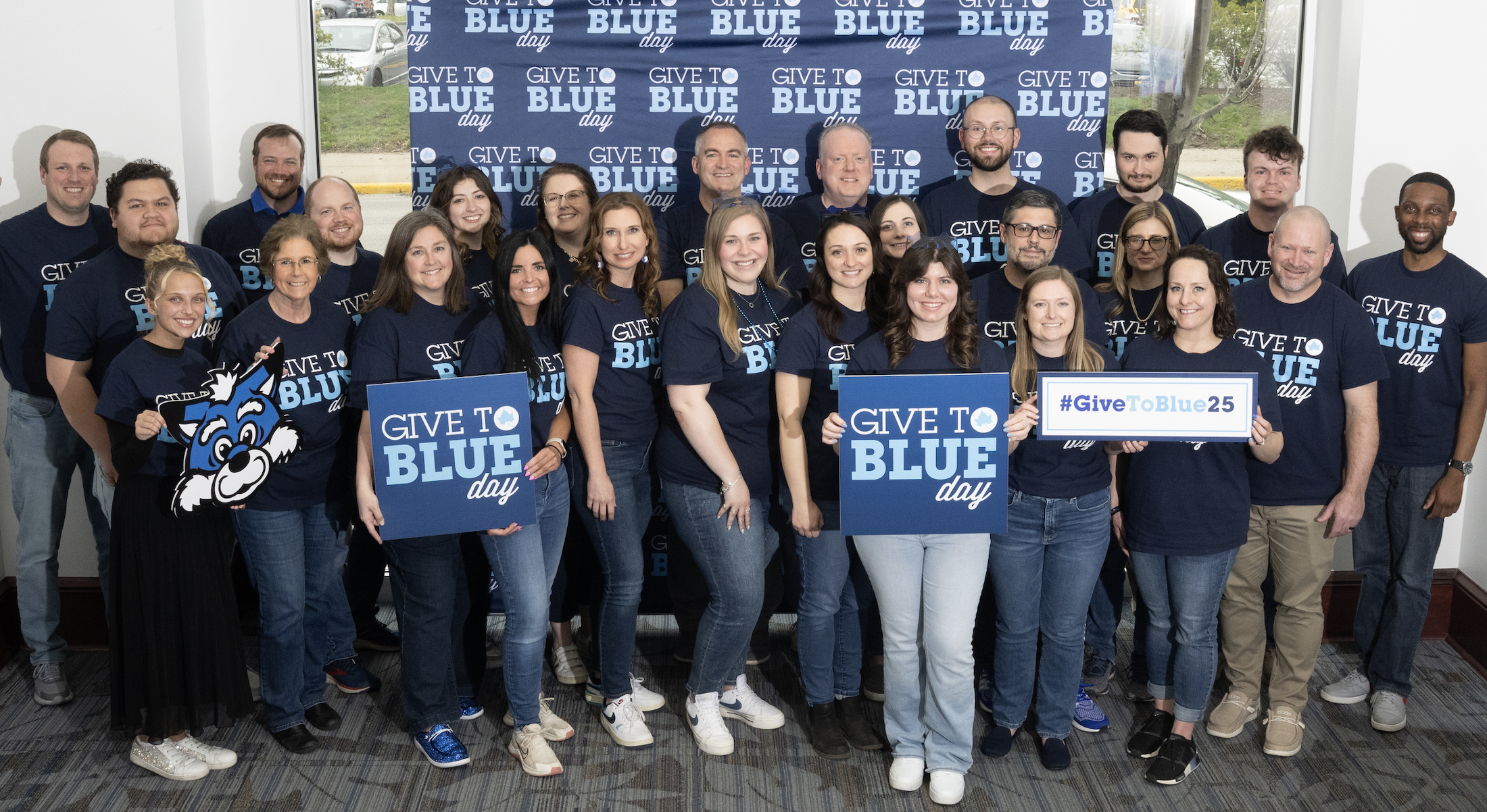 Group of diverse people wearing navy blue Give to Blue Day t-shirts, holding signs and mascot cutout, smiling in front of a Give to Blue Day backdrop.