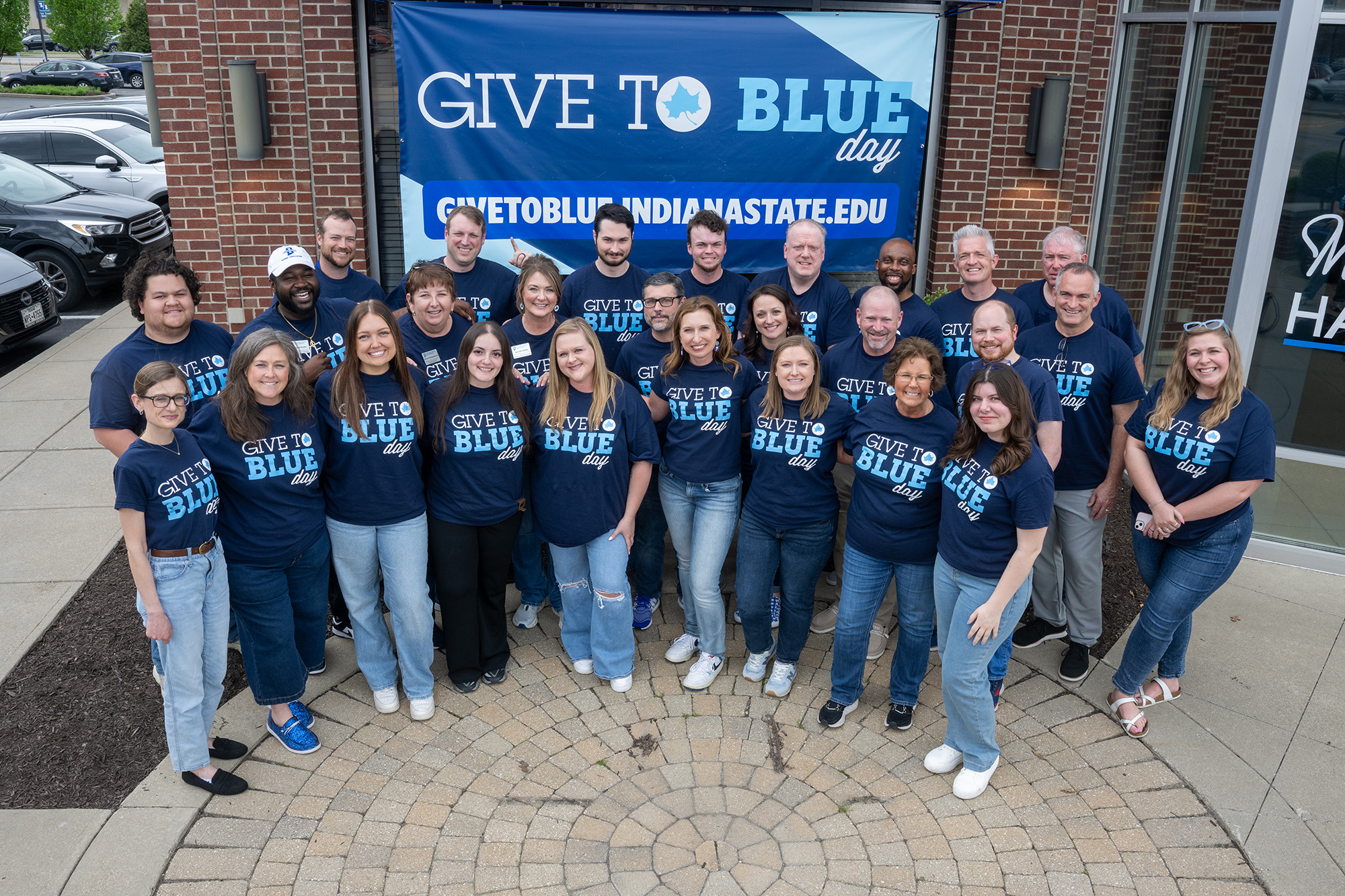 Group of 27 people smiling and wearing matching navy blue "Give to Blue day" t-shirts standing in front of a blue and white Give to Blue Day banner.