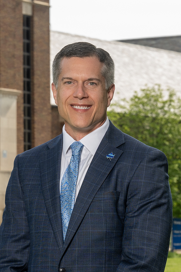 Man in a blue checkered suit and patterned blue tie smiling, standing outdoors with a building and trees in the background.