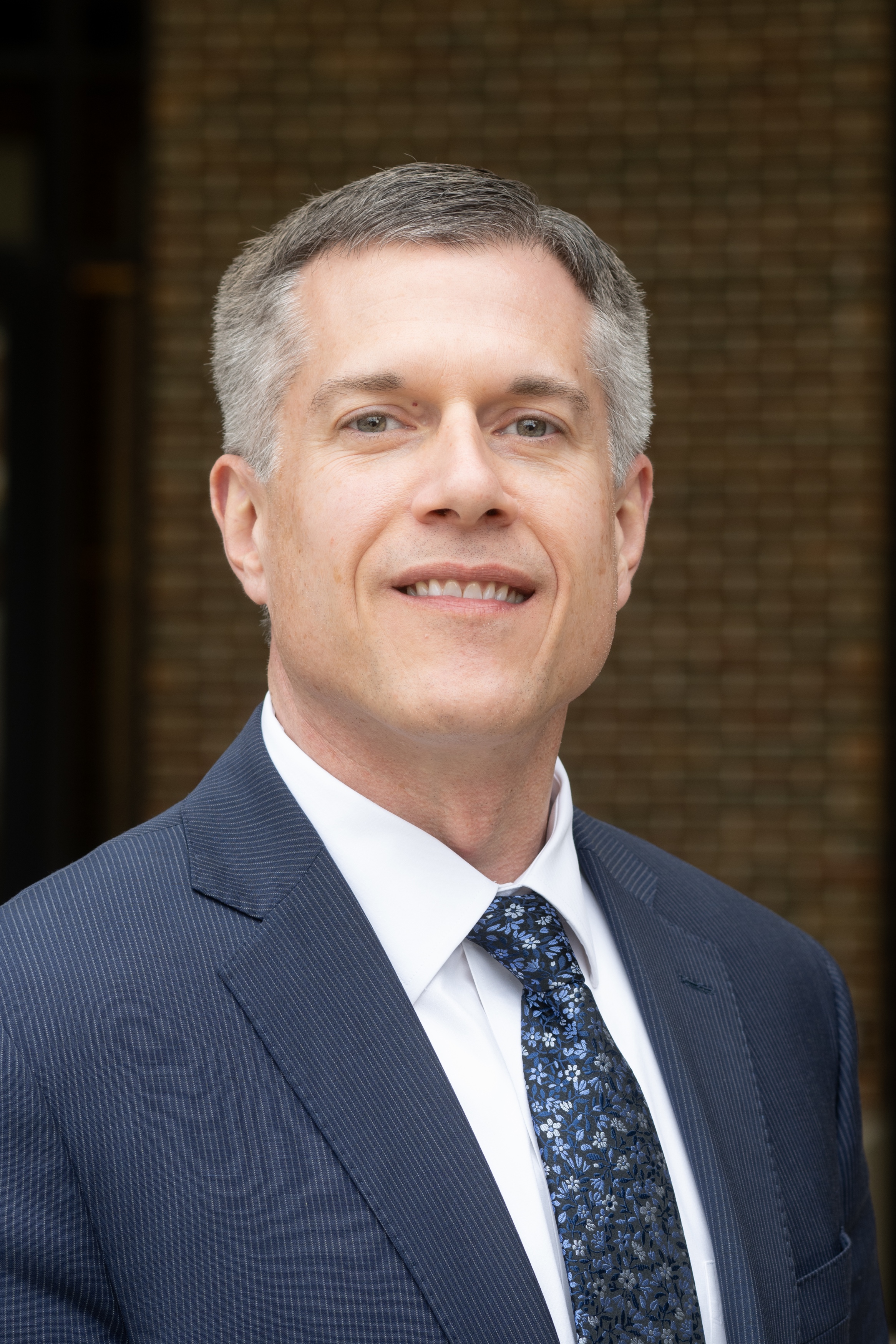 Middle-aged man with short salt-and-pepper hair wearing a pinstriped navy suit, white shirt, and floral-patterned dark tie, smiling slightly.