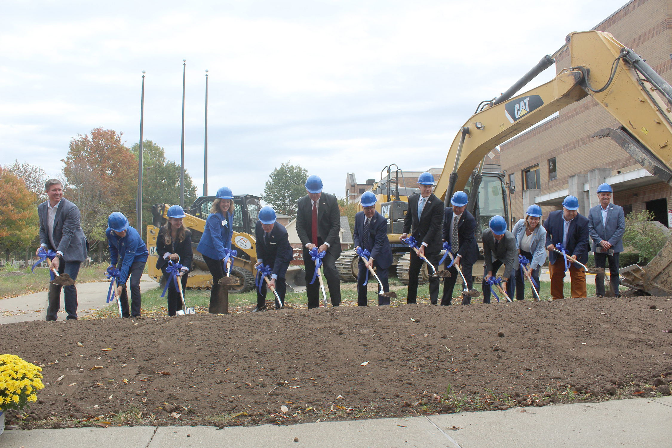 A group of thirteen people wearing blue hard hats breaks ground with shovels at a construction site in front of excavators and a building.