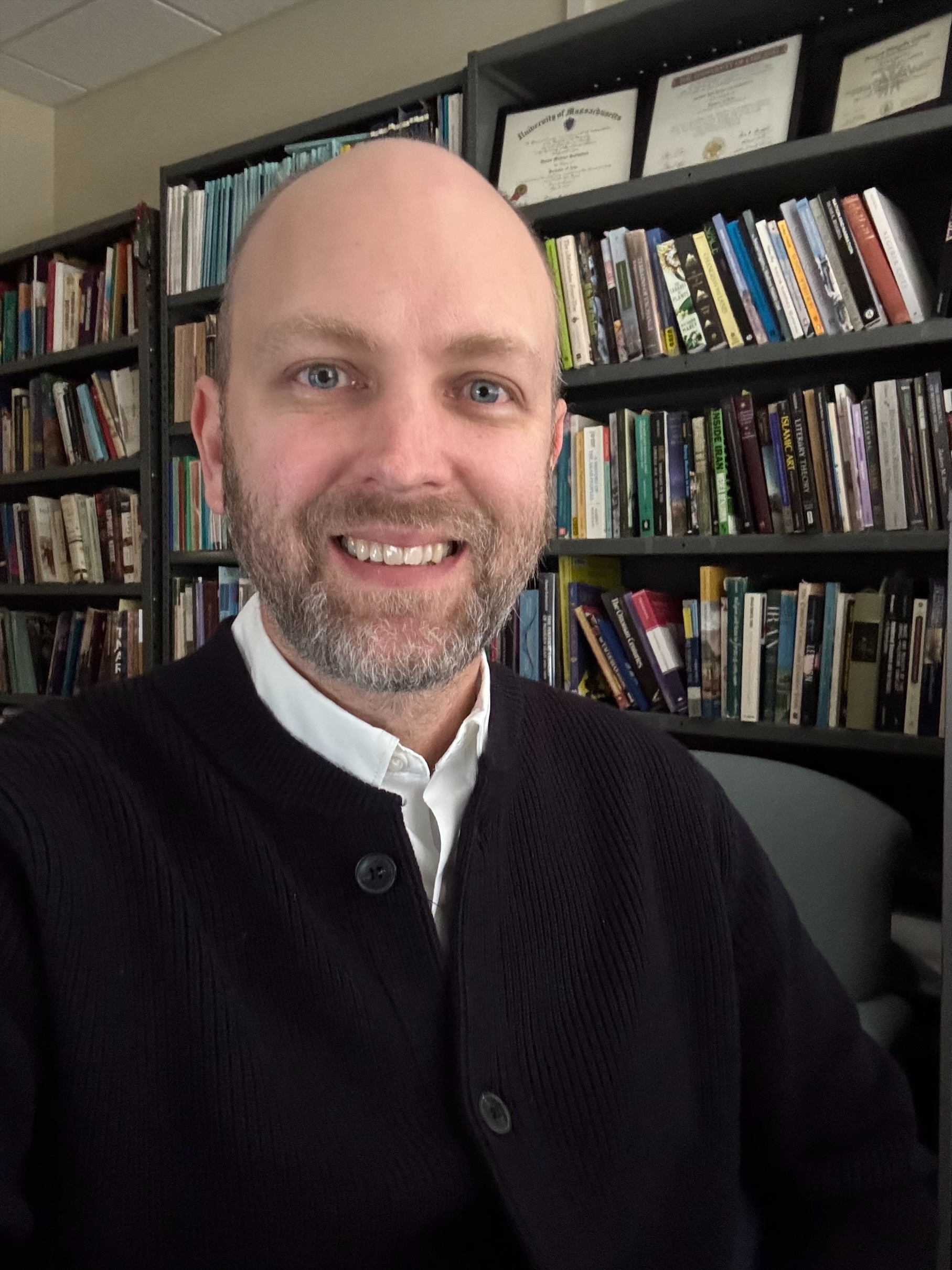 Smiling man with short beard and bald head wearing dark sweater and white shirt, sitting in front of bookshelves filled with books and framed certificates.