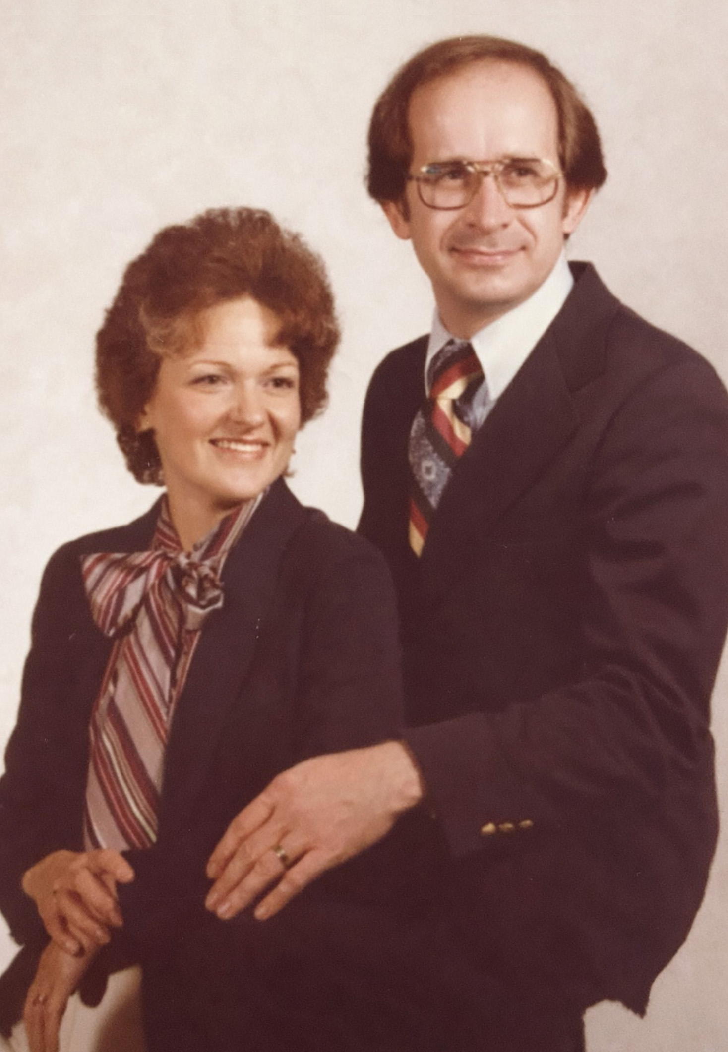 Middle-aged man and woman dressed formally, the man with glasses and a striped tie, the woman with curly hair and a bow-tie blouse, smiling and seated together.