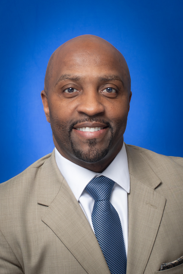 Smiling man with a shaved head and goatee wearing a tan suit jacket, white shirt, and blue striped tie against a blue background.