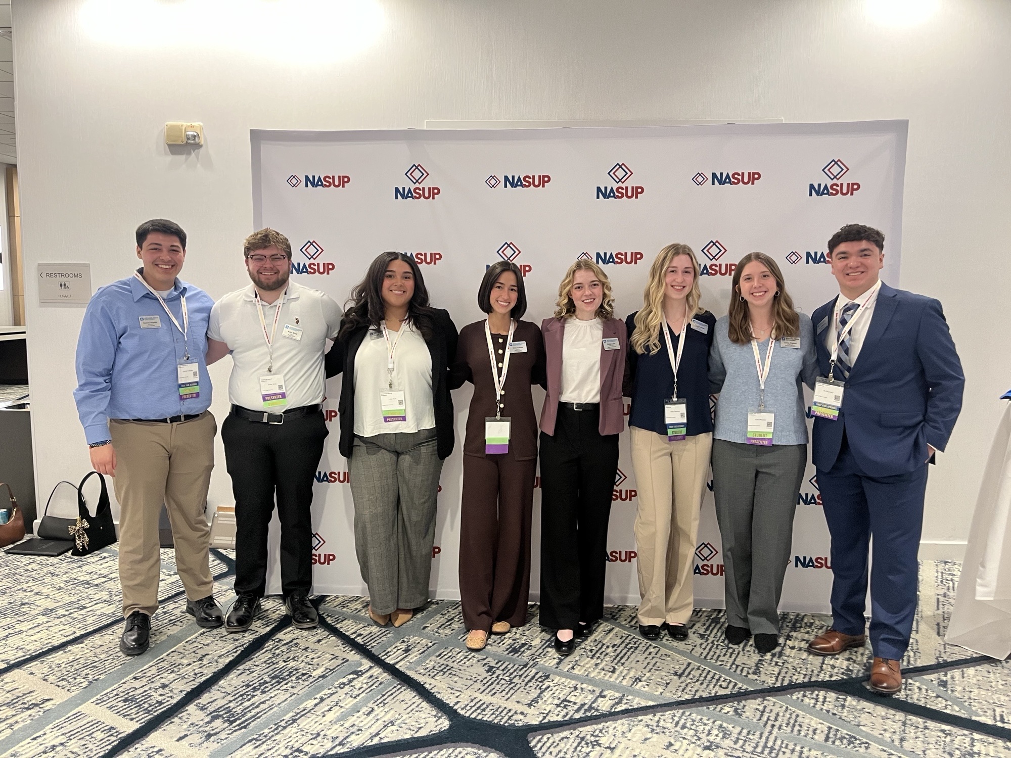 Eight young adults wearing conference badges stand shoulder to shoulder in front of a NASUP backdrop inside a conference venue.