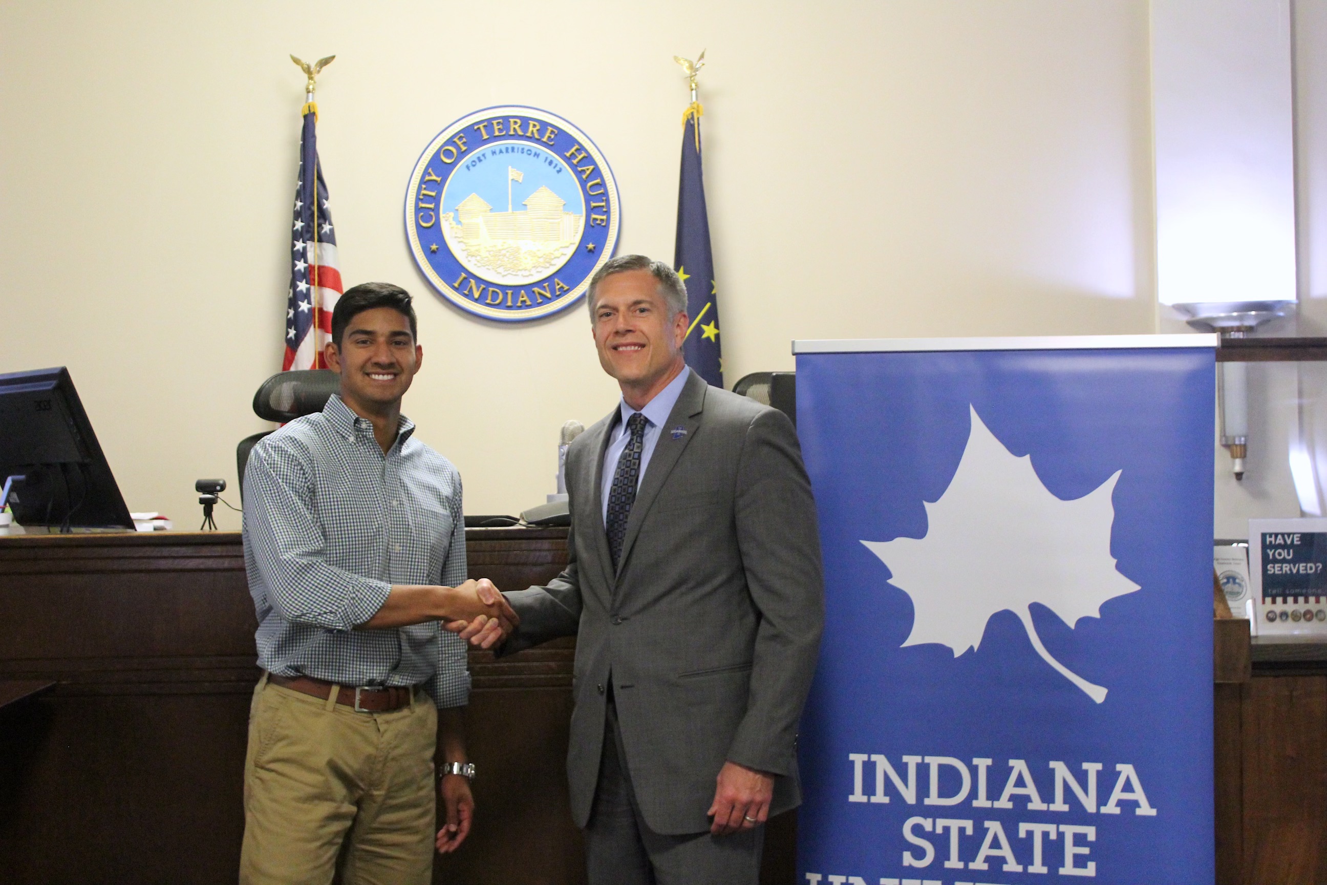 Two men shaking hands in a government office, city seal of Terre Haute and Indiana state flag behind, Indiana State University banner to right.