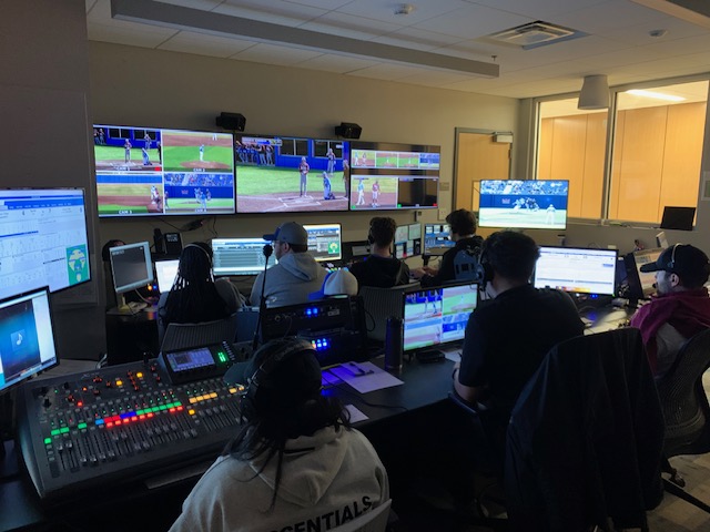 Broadcast production team at work in control room with multiple monitors showing baseball games and audio mixing equipment.