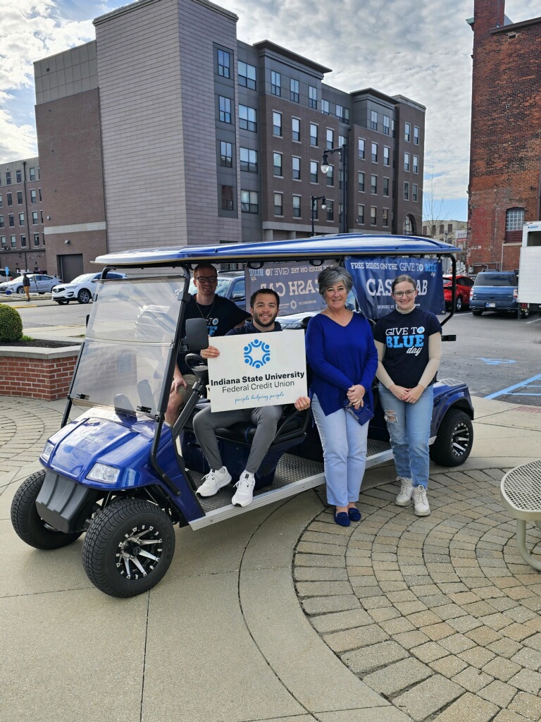 Four people smiling near and sitting on a blue golf cart in an urban plaza, one holding a sign for Indiana State University Federal Credit Union.