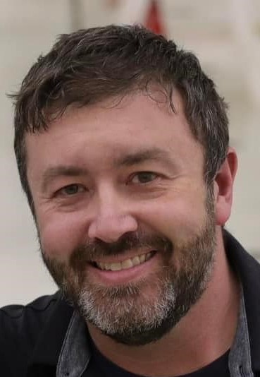 Smiling man with short dark hair and beard wearing a dark collared shirt in front of a blurred background.