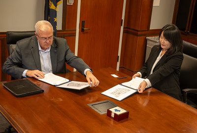 Two people seated at a conference table signing documents during a formal meeting in a wood-paneled room.
