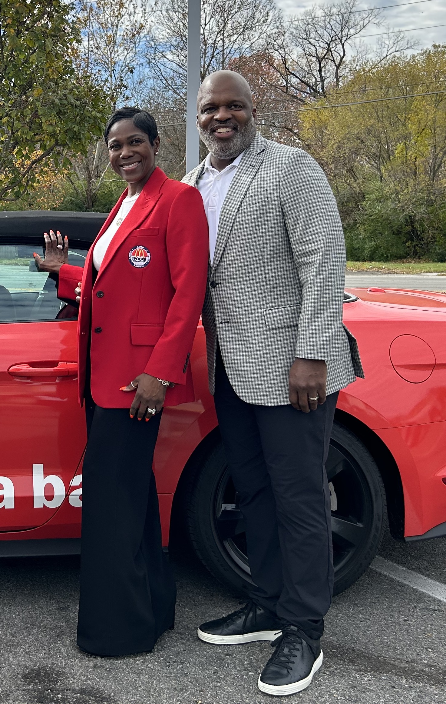 Two people smiling and standing beside a red car, outdoors with trees in the background. One person wears a red blazer with a badge, the other a checked blazer.