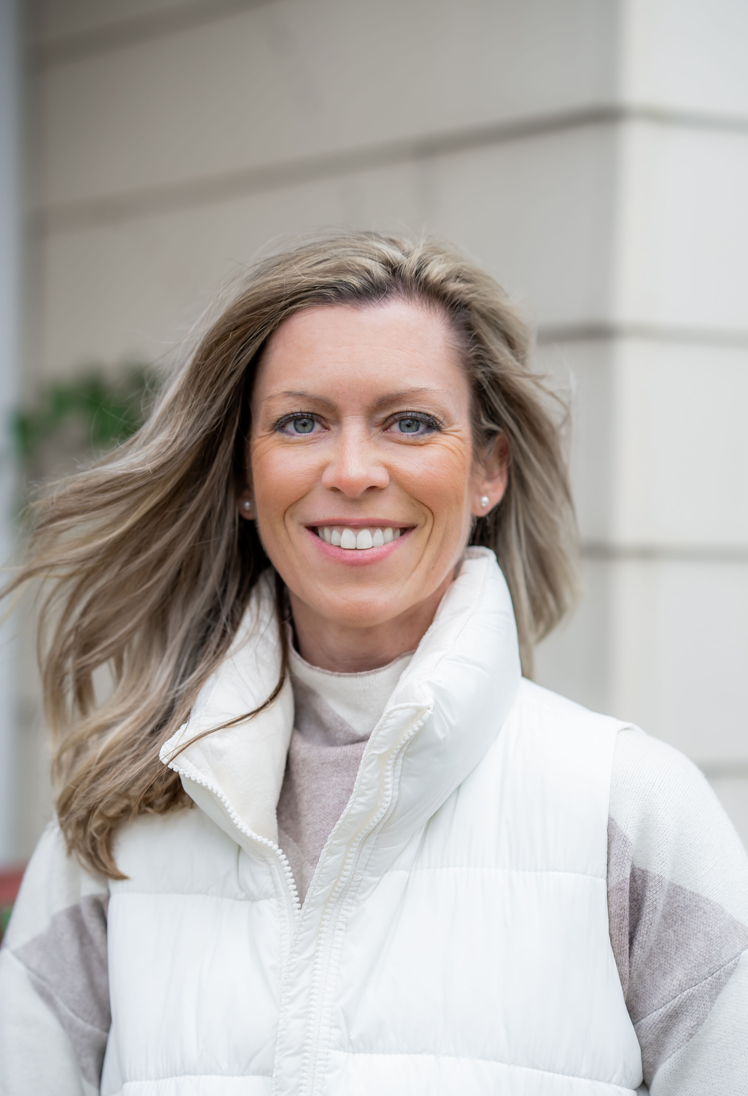Smiling woman with light brown hair wearing a white puffer vest and striped sweater, standing outdoors near a light-colored building wall.