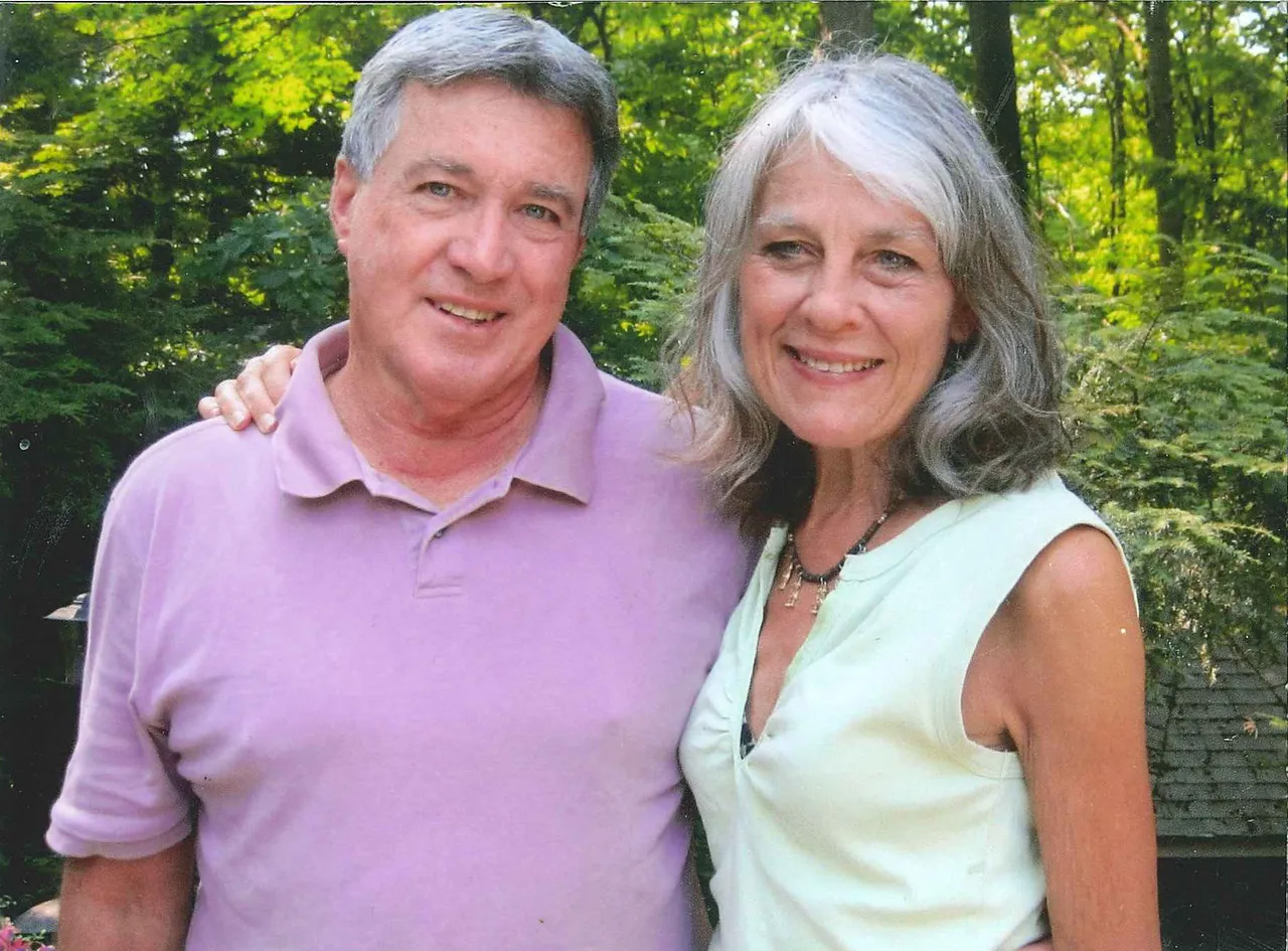 Middle-aged man in a light purple polo shirt stands with arm around a woman with gray hair wearing a sleeveless light green top, outdoors with trees.