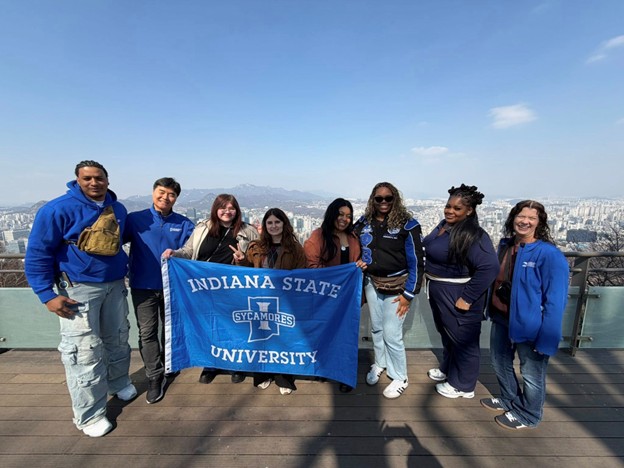 Seven people standing outdoors on a rooftop with a cityscape and mountains in the background, holding an Indiana State University banner.