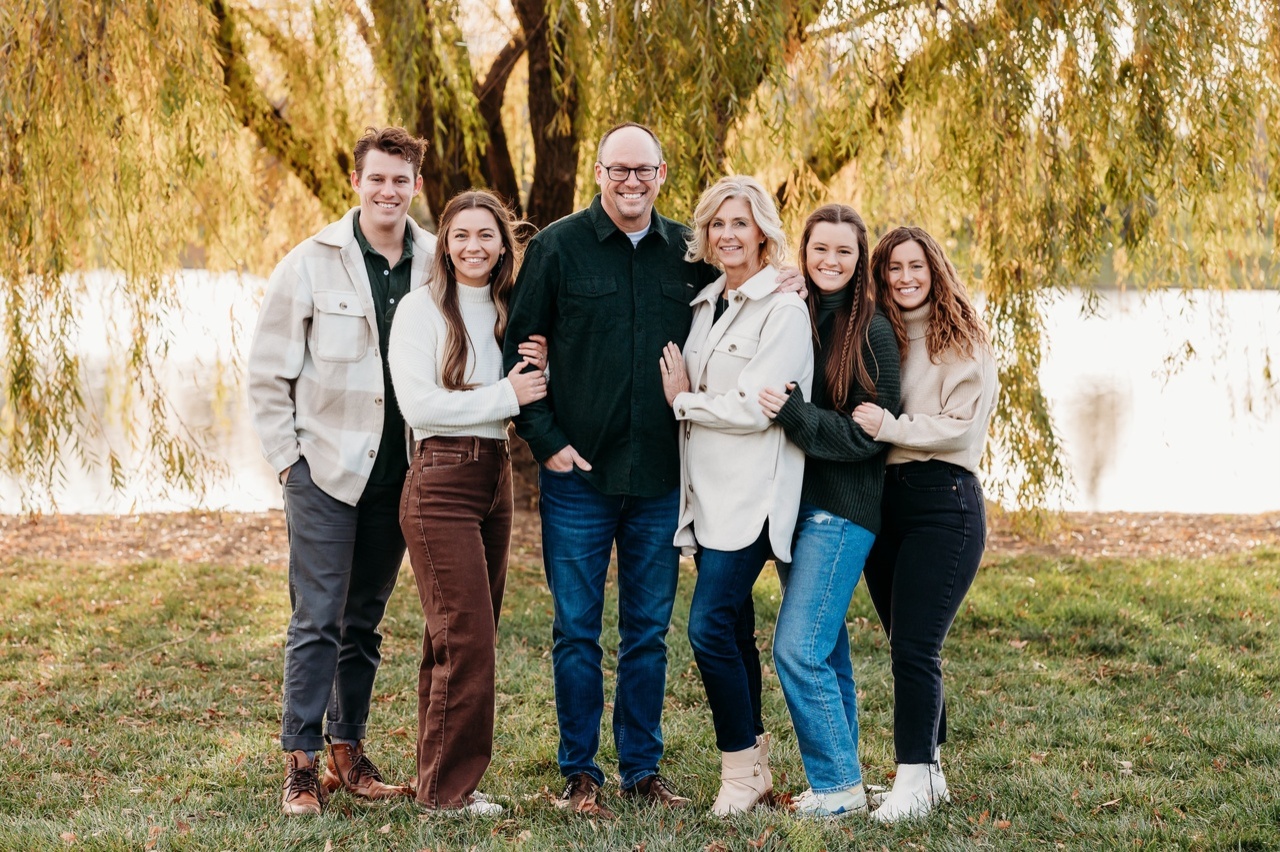 Six people standing closely together outdoors in front of a tree with hanging branches near a body of water, smiling at the camera.