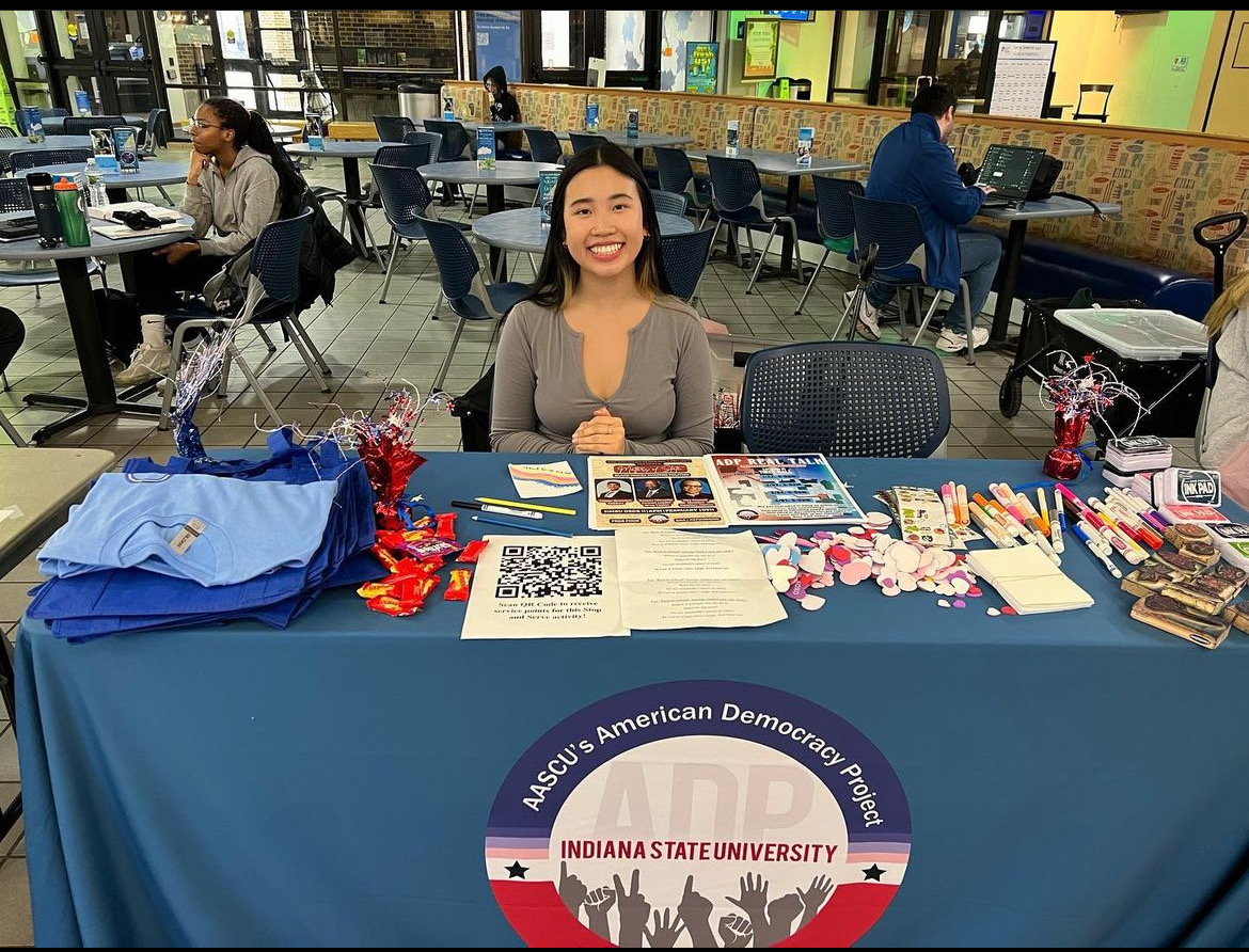 Young woman sitting at a table with promotional materials and giveaways for Indiana State University’s American Democracy Project in a campus common area.
