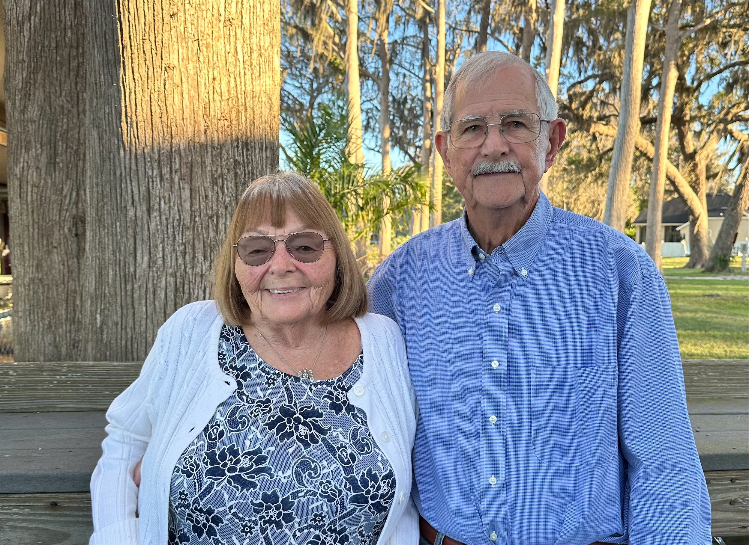 Elderly man and woman standing outdoors in front of trees, both wearing glasses; woman in a floral dress and white cardigan, man in a blue button-up shirt.