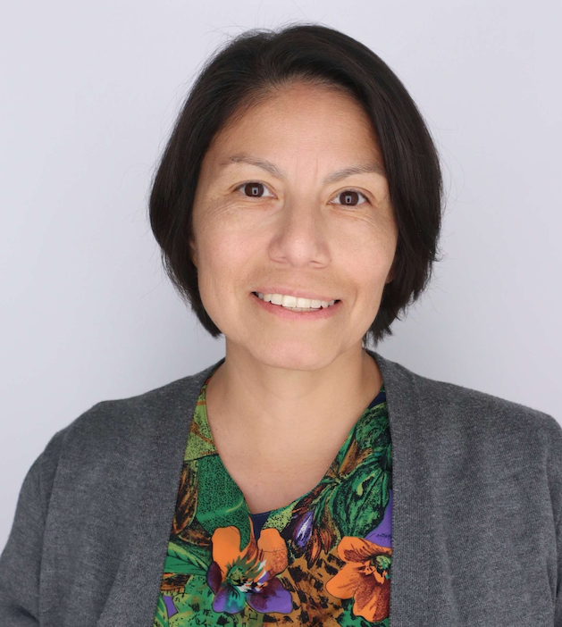 Smiling woman with short dark hair wearing a gray cardigan over a colorful floral top against a plain white background.