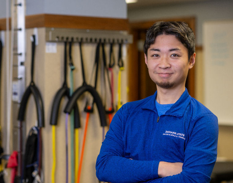 Young man in blue Indiana State University athletic training jacket stands with arms crossed in front of resistance bands and exercise equipment.