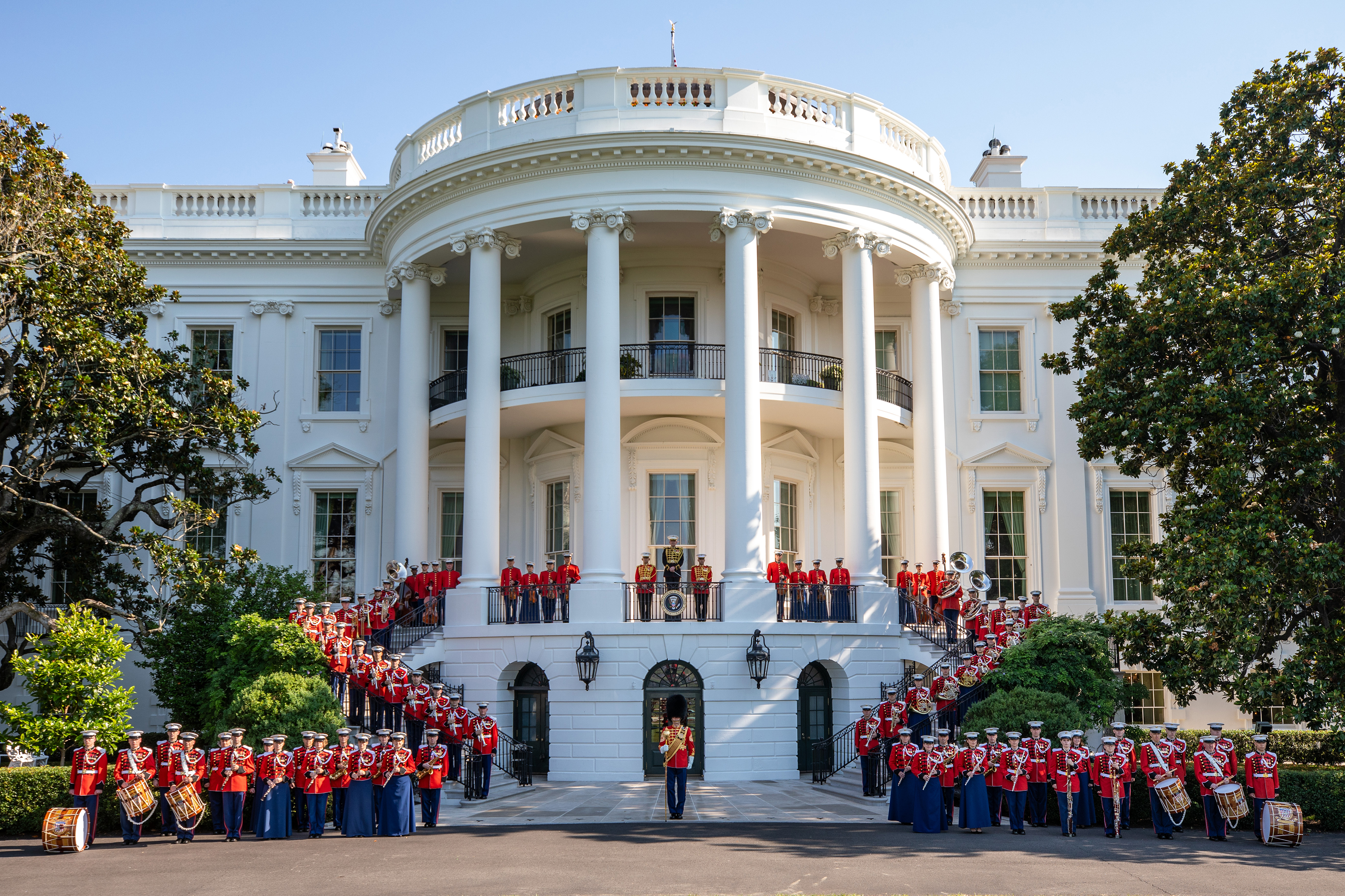 Large United States Marine Band in red uniforms posed in front of the White House South Portico with instruments and drums.