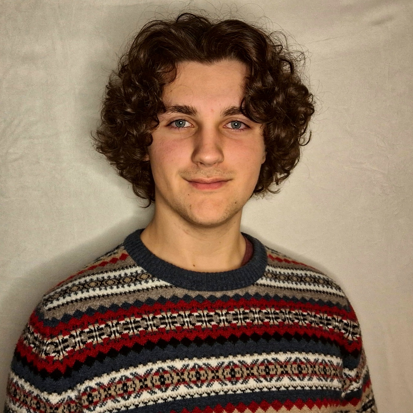 Young person with curly brown hair wearing a patterned sweater with red, navy, beige, and white colors, standing against a plain light-colored background.