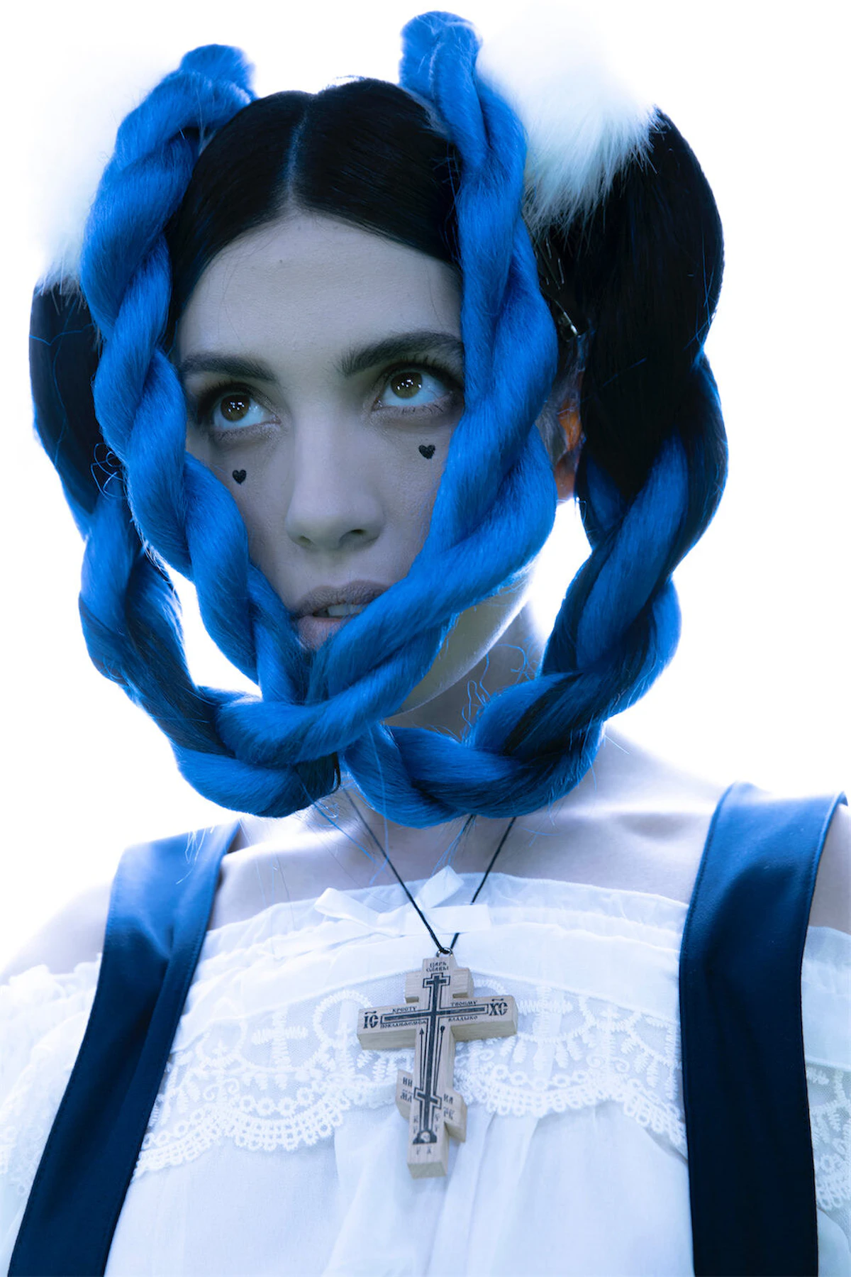 Woman with large braided blue hair styled around her face, wearing a white lace top, black strap, and a wooden cross necklace, with small black heart face tatto