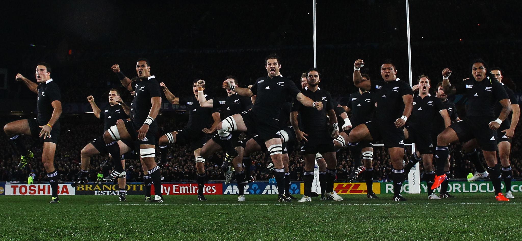 Rugby players in black uniforms performing the Haka, a traditional Māori dance, on a stadium field with goalposts and a crowd in the background.