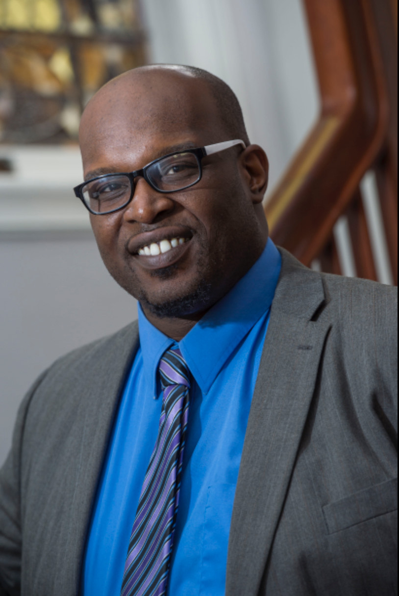 Man wearing glasses, a blue shirt, striped tie, and gray blazer smiling indoors near a staircase.