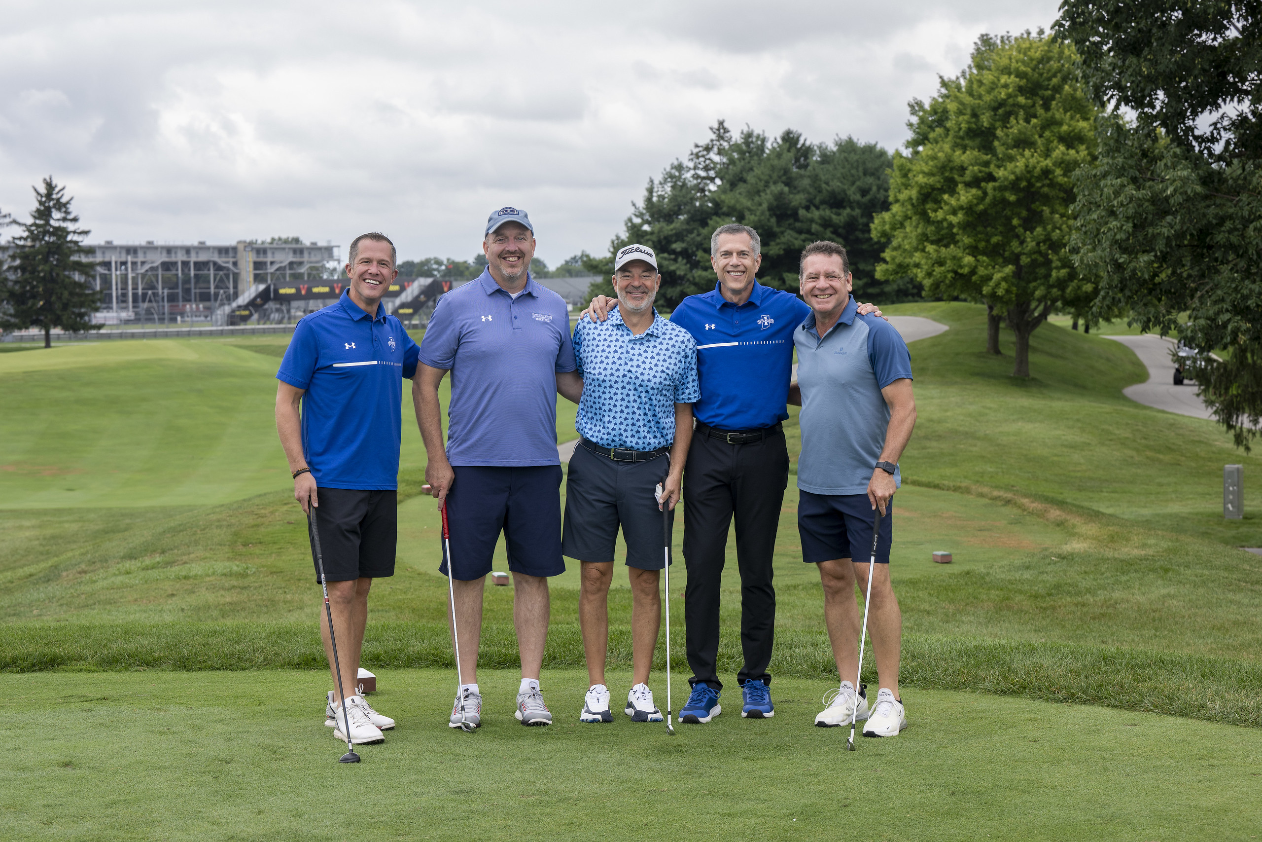 Five men standing on a golf course fairway, smiling, dressed in golf attire, each holding a golf club, with trees and a stadium in the background.