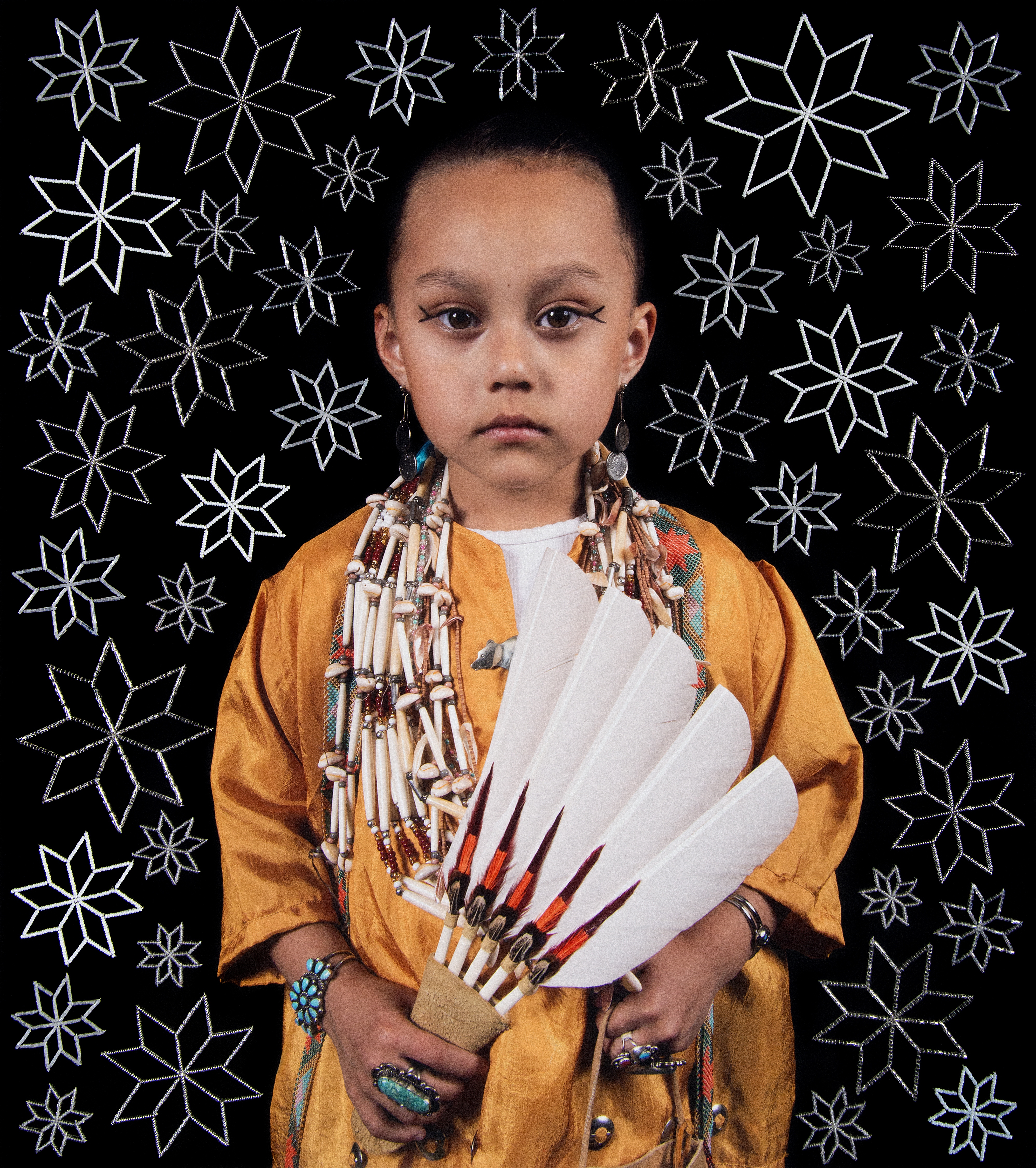 Young Indigenous child in traditional orange garment holds a fan of white feathers with red tips, wearing bead jewelry, against a black background with white st