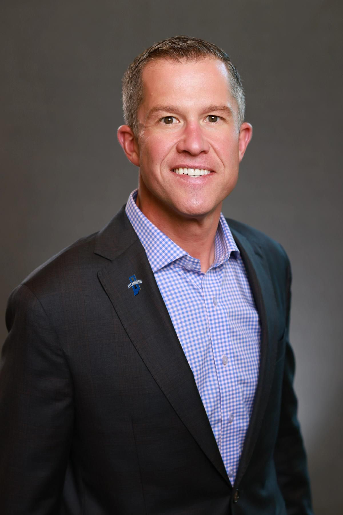 Smiling man in a dark suit jacket with a blue checkered shirt, wearing a small Indiana State Sycamores pin on the lapel, gray background.