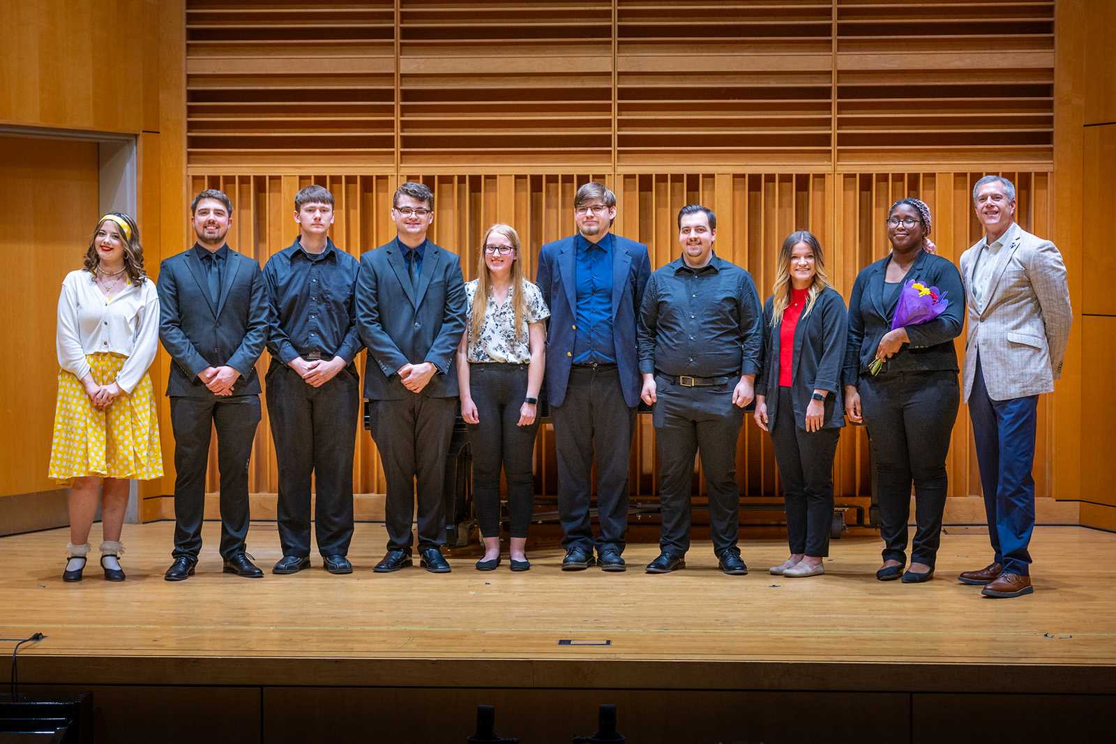 Ten young adults and one older man standing on a wooden stage in a concert hall, one woman holding a bouquet of flowers.