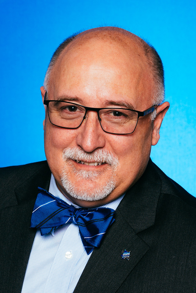 Bald man with glasses, gray goatee, wearing a blue striped bow tie, light blue shirt, dark suit jacket, and a star-shaped pin on lapel, blue background.