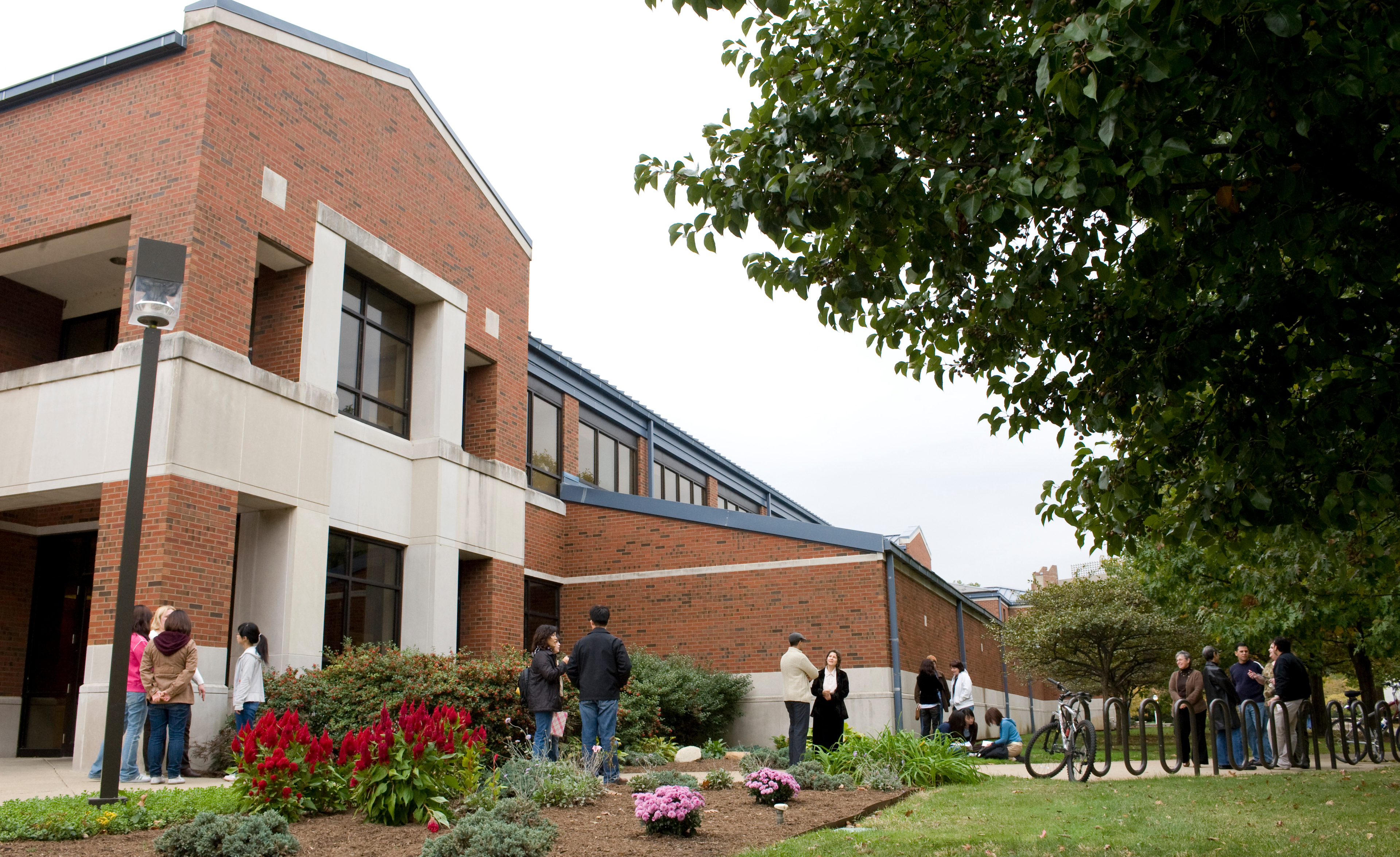 Groups of people standing and talking on a college campus near a brick building with flowers and bike racks under a tree canopy.