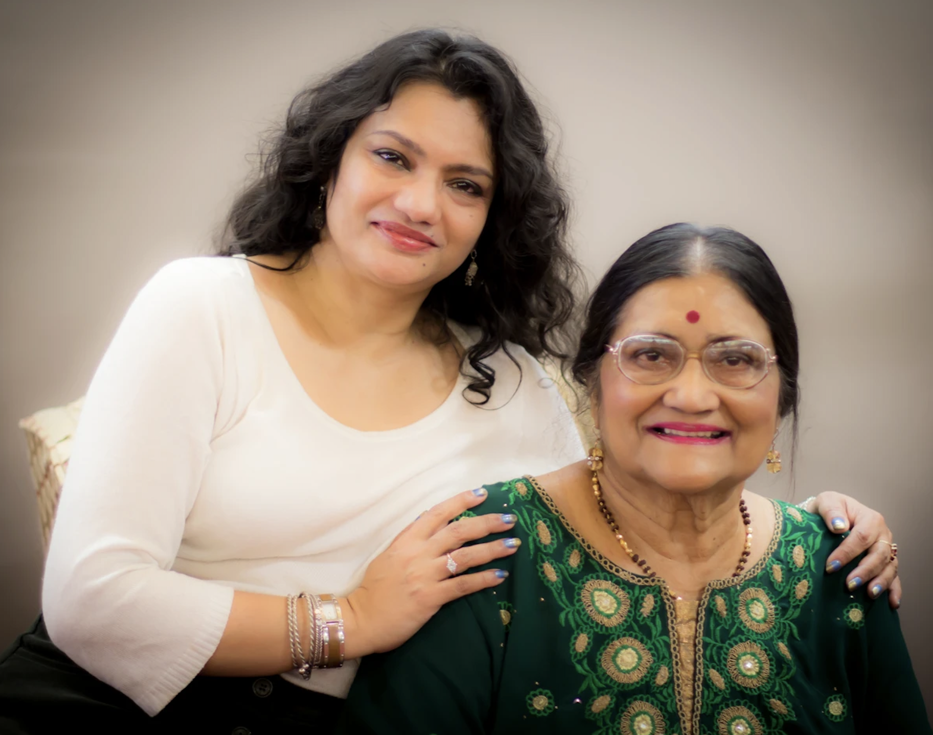Two women smiling, one younger with wavy hair and white top resting hand on the shoulder of an older woman wearing glasses and a green embroidered outfit.