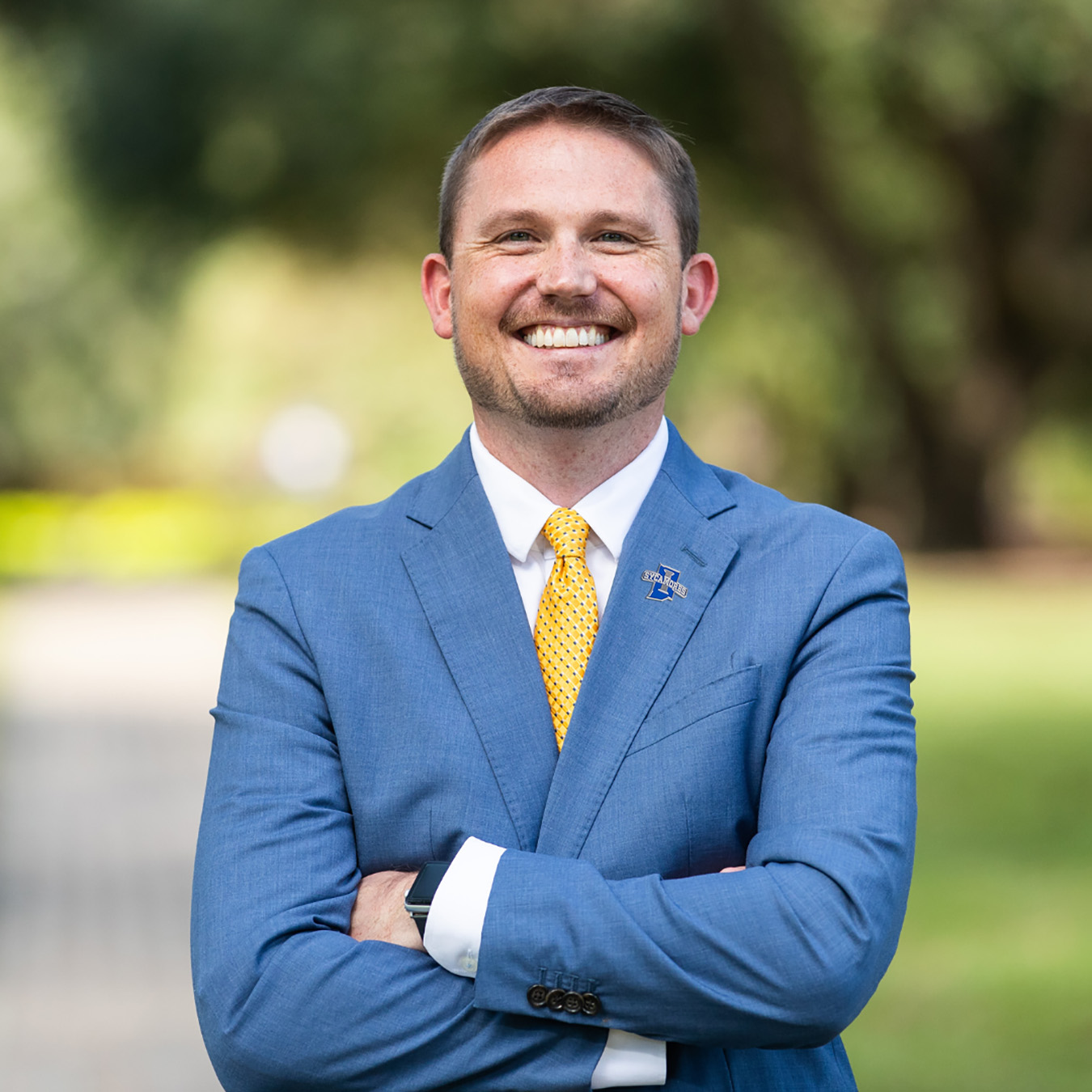 Smiling man in blue suit, white shirt, and yellow tie standing outdoors with arms crossed, wearing a lapel pin with "Sycamores" logo.