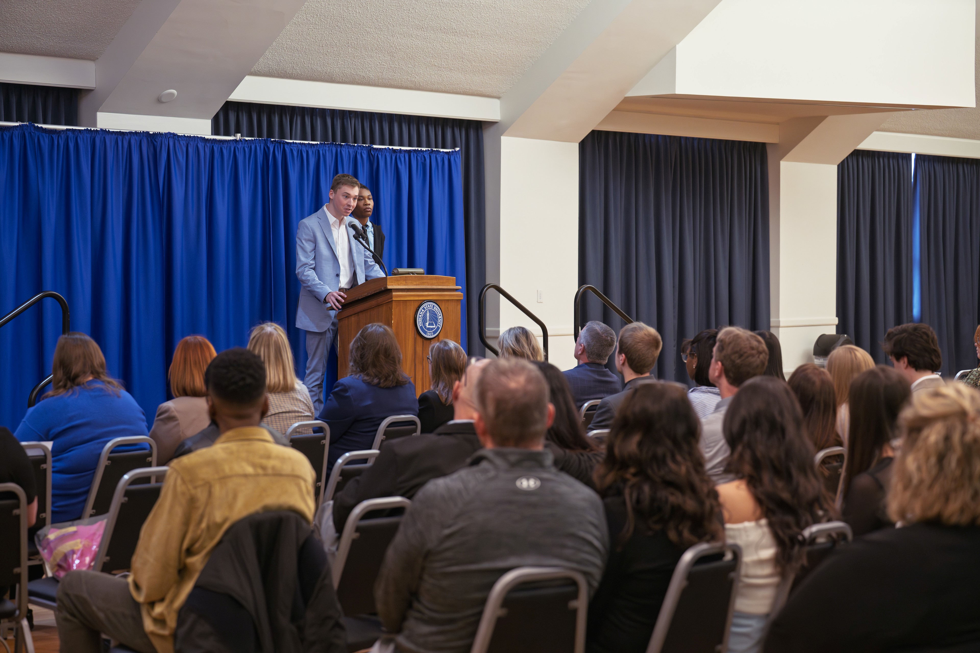 Young man and woman speaking at a podium in front of an audience seated facing them, blue curtain backdrop, indoor event setting.