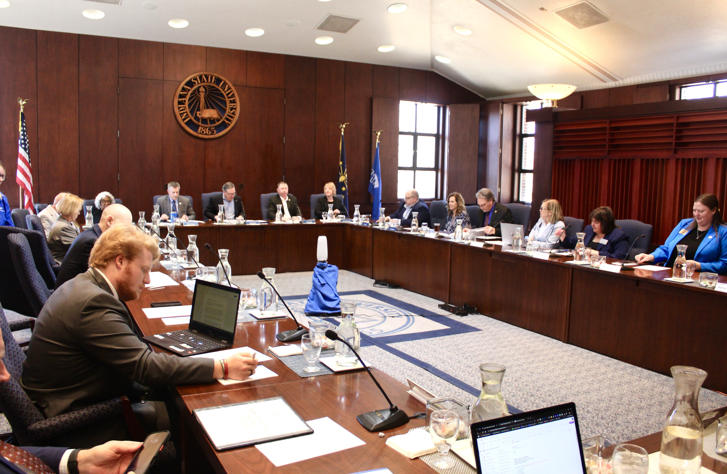 University board members seated around a large U-shaped wood conference table during a meeting in a room with Indiana State University emblem on the wall.